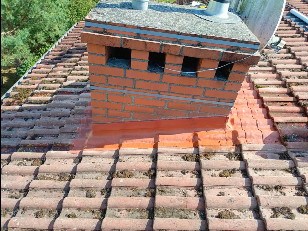 Ceglany komin z otworami wentylacyjnymi na dachu z dachówki ceramicznej, z widocznymi śladami mchu i zabrudzeń. Na kominie antena satelitarna.