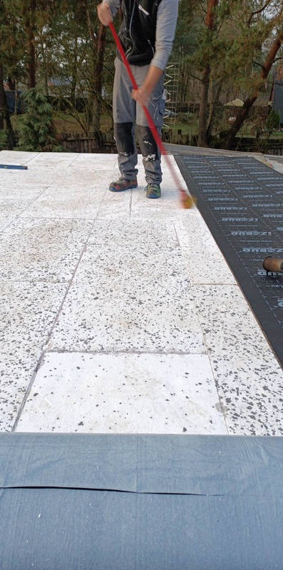 Pracownik rozprowadza masę bitumiczną na płaskim dachu za pomocą czerwonego wałka. Część dachu pokryta jest już czarną membraną izolacyjną.