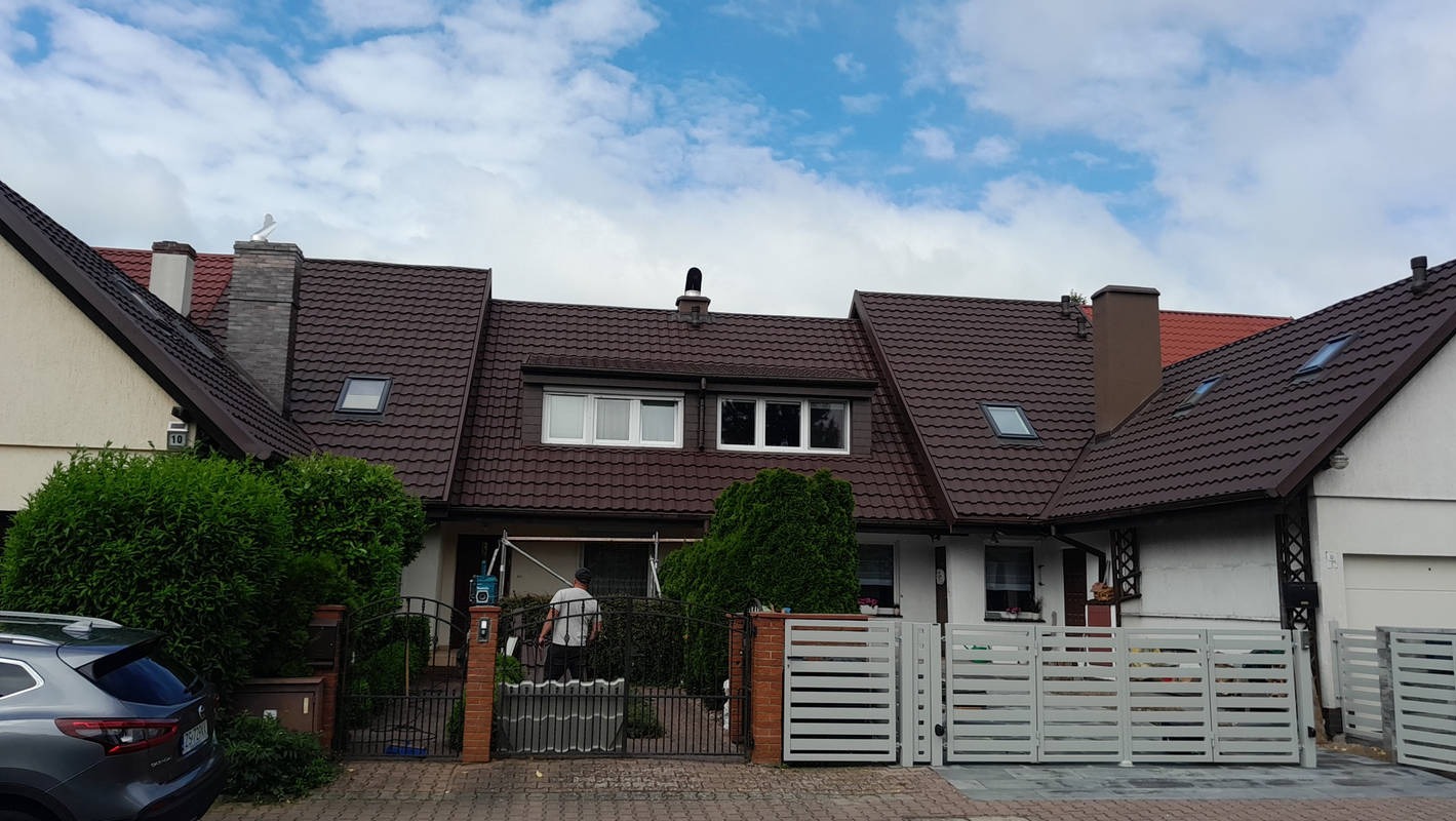 Rząd domów z brązowymi dachami z dachówki, barierki w kolorze białym. Na jednym z kominów ozdobna figurka ptaka. Osoba pracuje przy elewacji jednego z budynków.