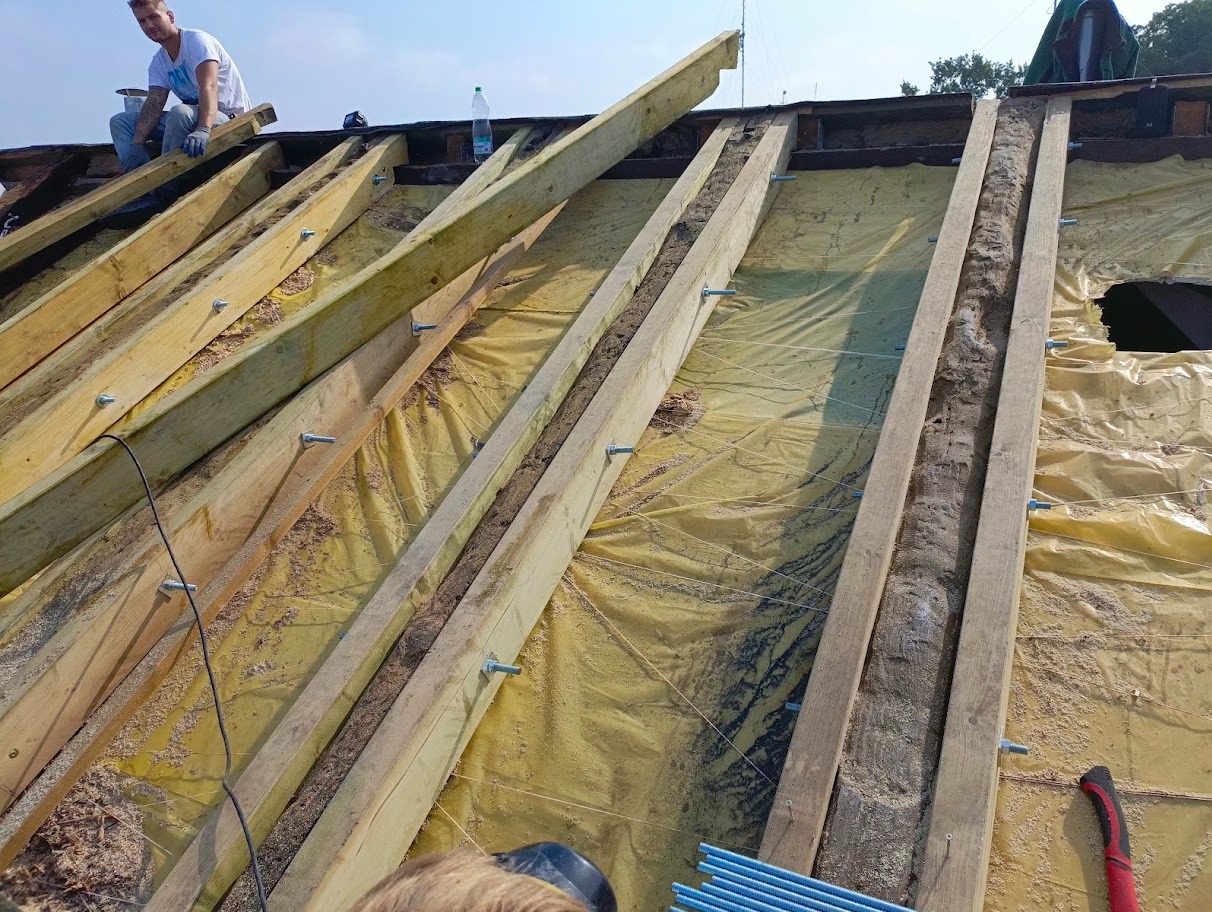 Konstrukcja dachu w trakcie naprawy, widoczne drewniane belki mocowane śrubami, żółta folia izolacyjna i pracownik w białej koszulce.