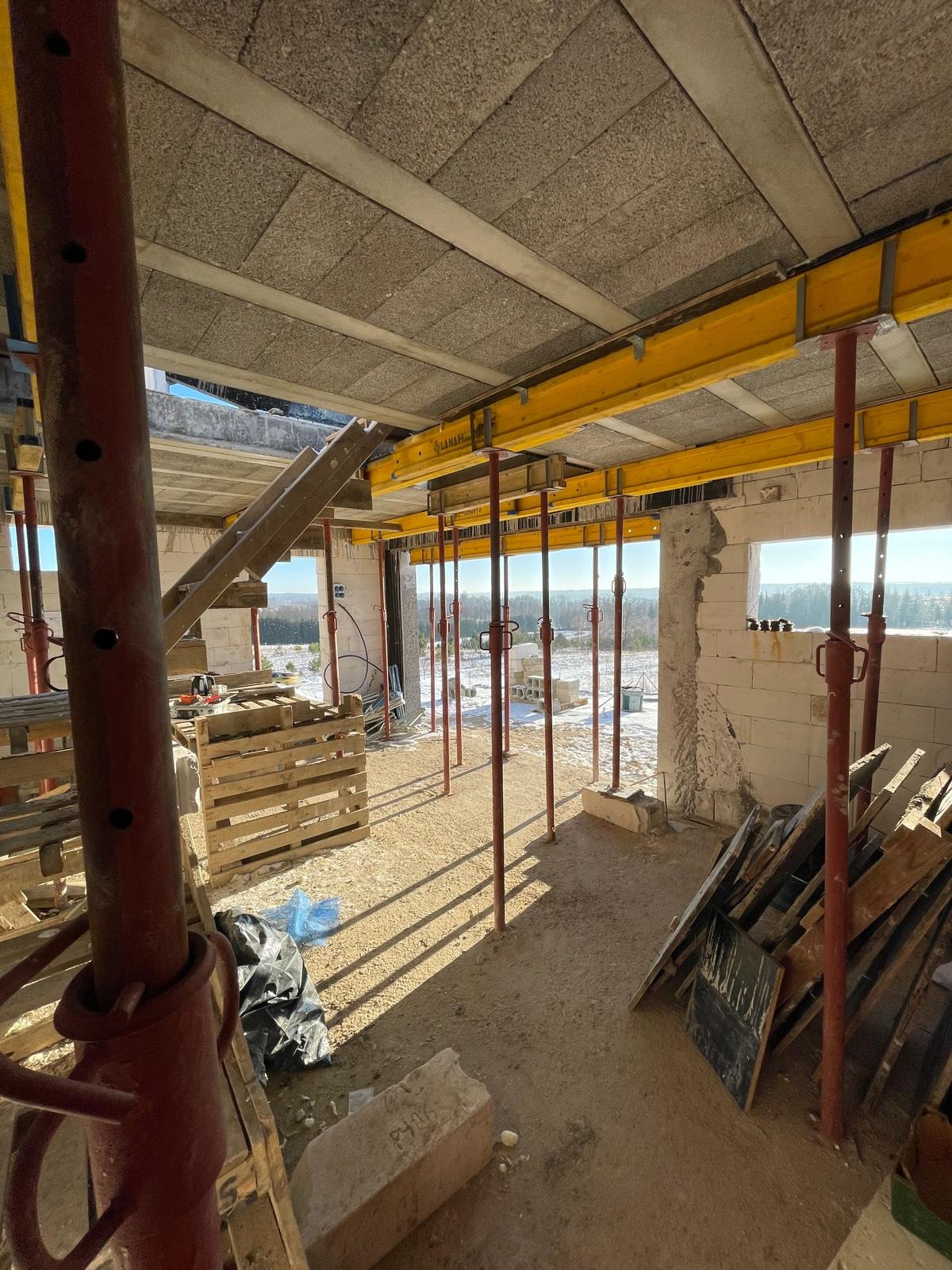 Wnętrze surowego domu w budowie: widoczne podpory stropu, bloczki, palety i narzędzia. W tle zimowy krajobraz za oknem. Etap prac murarskich.