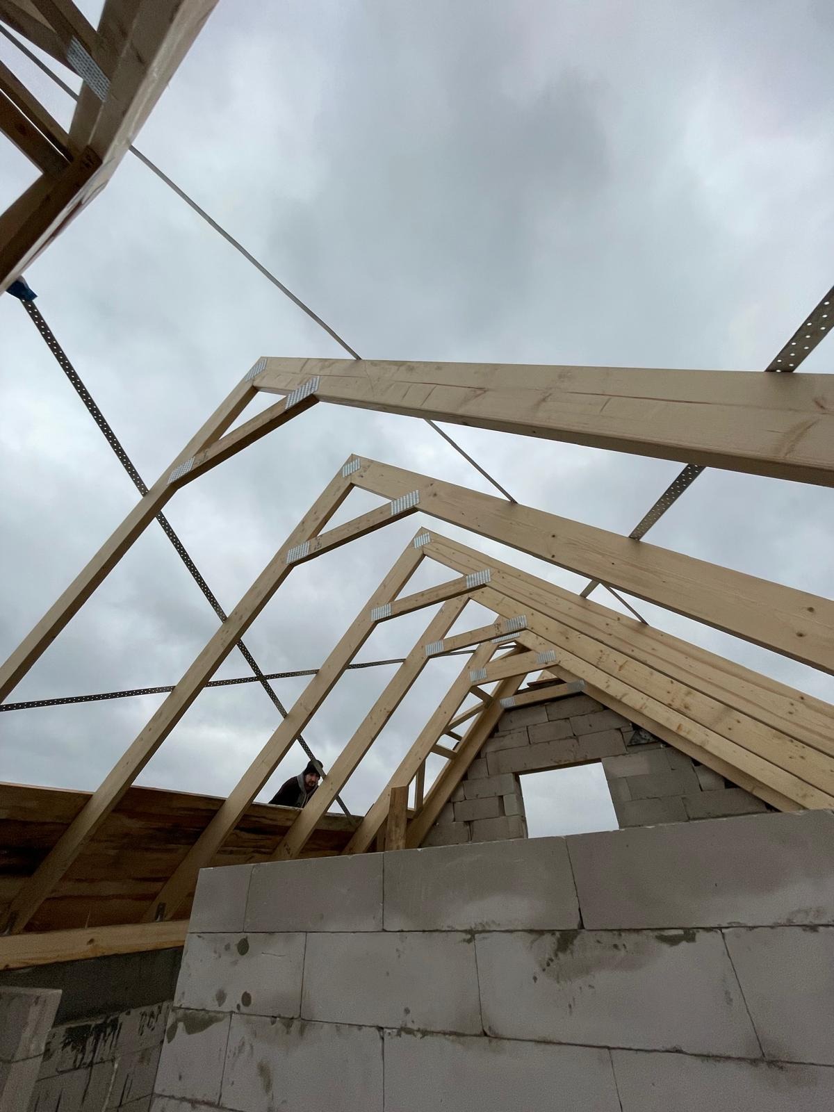 Konstrukcja drewniana dachu w trakcie budowy, widok od dołu na szkielet z belek i metalowych łączników na tle pochmurnego nieba. Mur z bloczków.