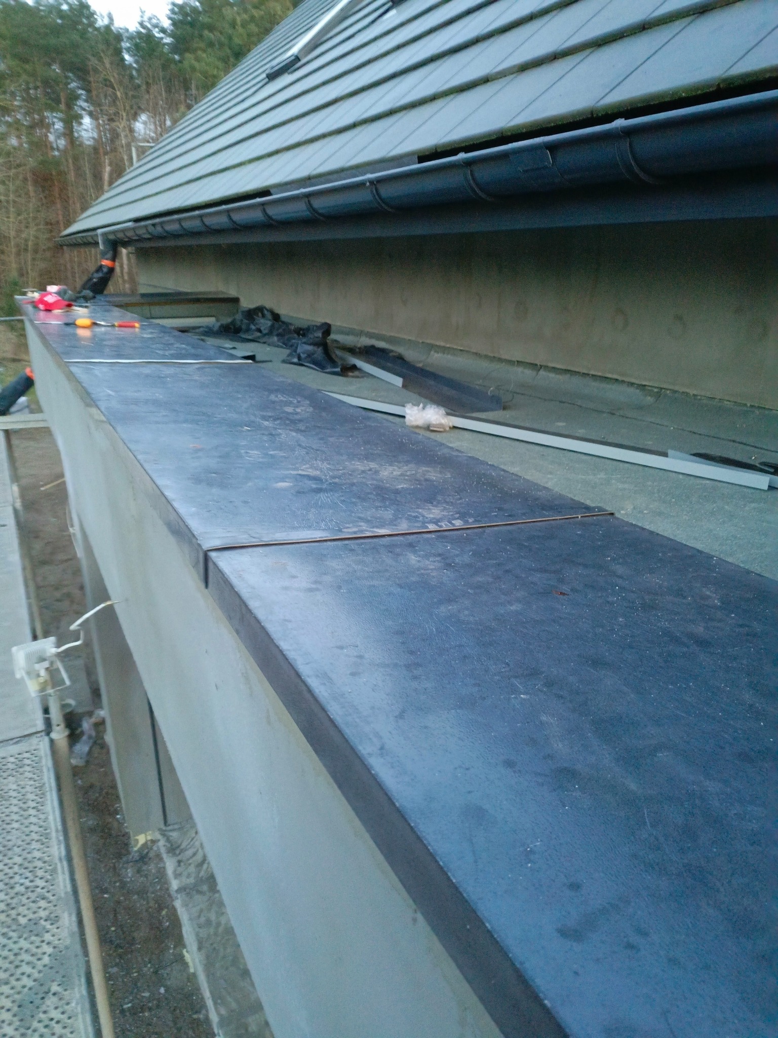 Montaż obróbek blacharskich na budynku z szarą dachówką. Widoczne narzędzia i elementy instalacji. Detal wykończenia elewacji w trakcie prac.