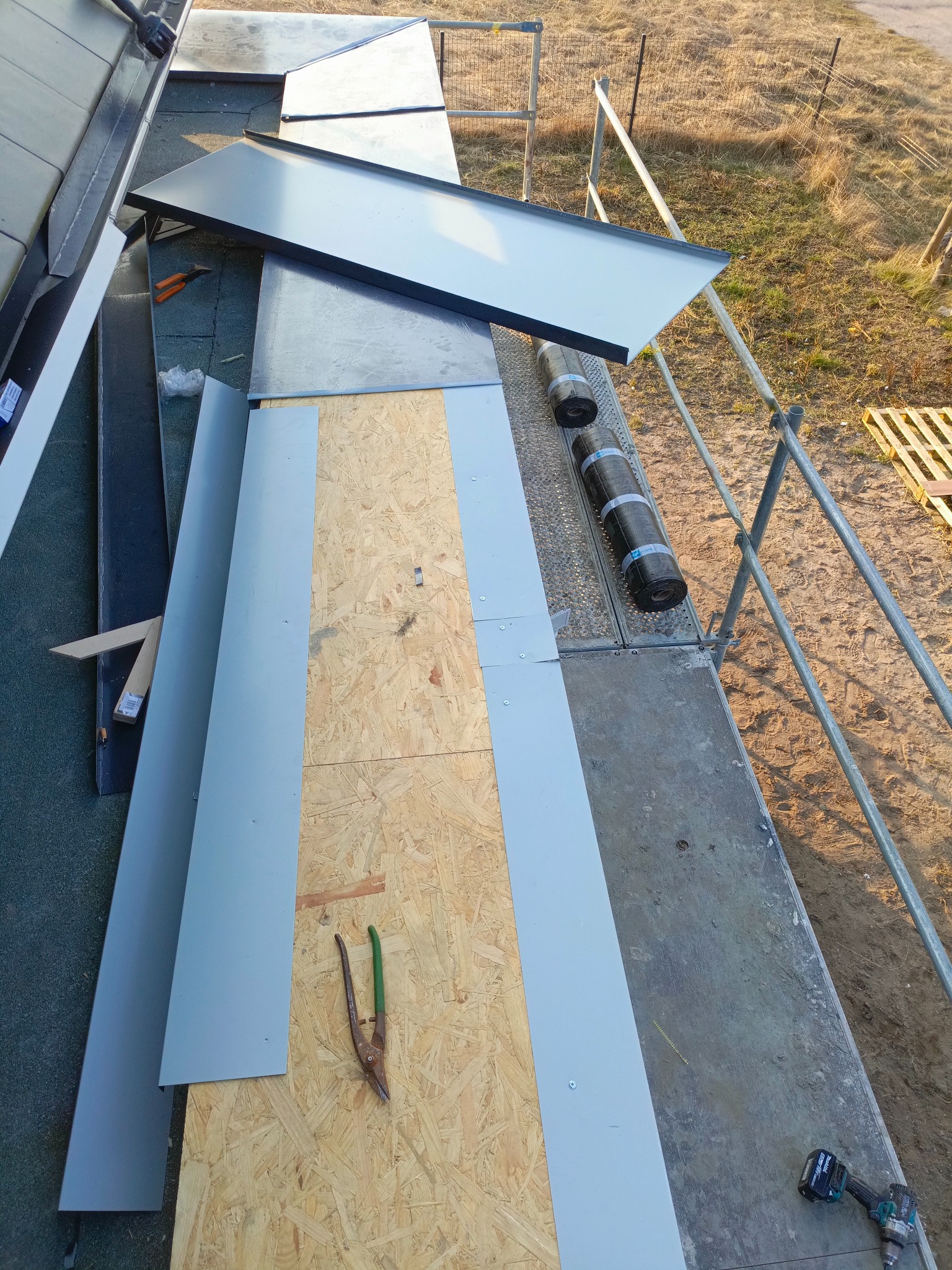 Montaż blaszanych elementów na dachu z widocznymi narzędziami dekarskimi i rolkami papy na rusztowaniu. Prace dekarskie w toku, widok z góry.