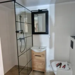 Montaż kabiny prysznicowej z czarnymi profilami i drewnianej szafki pod umywalkę w łazience z białymi płytkami, widoczne narzędzia i akcesoria instalacyjne na podłodze imitującej drewno.