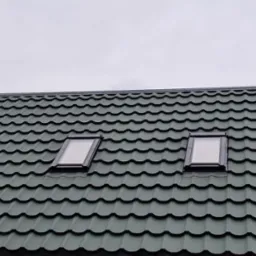 Zielony dach z blachodachówki z dwoma oknami dachowymi, widok z dołu na tle pochmurnego nieba.