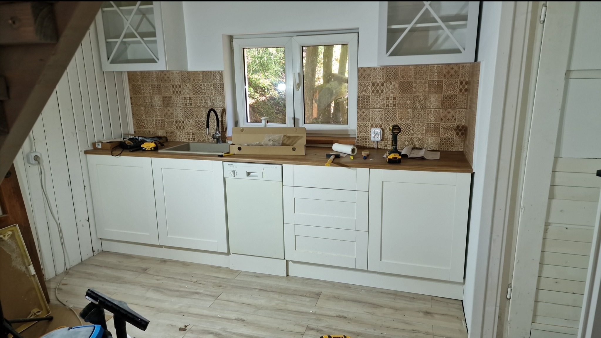 Montaż białej zabudowy kuchennej z drewnianym blatem i wzorzystymi płytkami backsplash, widoczne narzędzia i akcesoria montażowe.