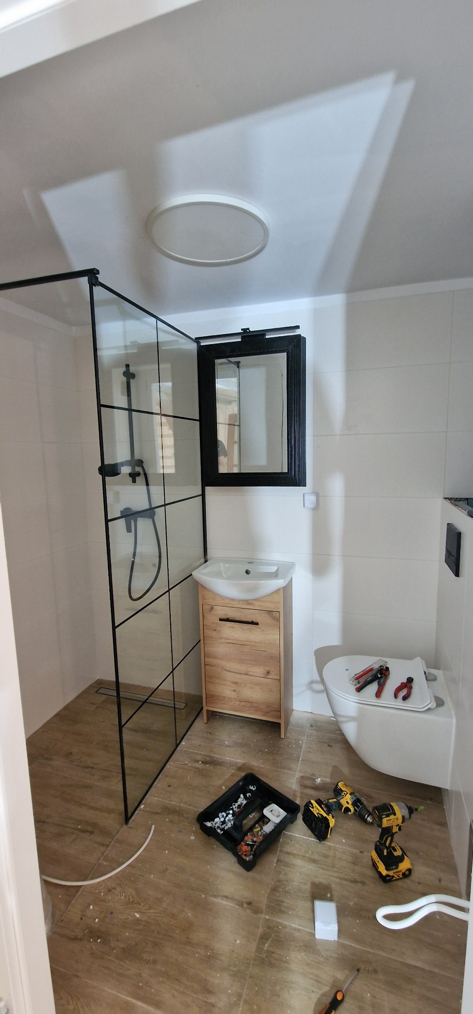 Montaż kabiny prysznicowej z czarnymi profilami i drewnianej szafki pod umywalkę w łazience z białymi płytkami, widoczne narzędzia i akcesoria instalacyjne na podłodze imitującej drewno.
