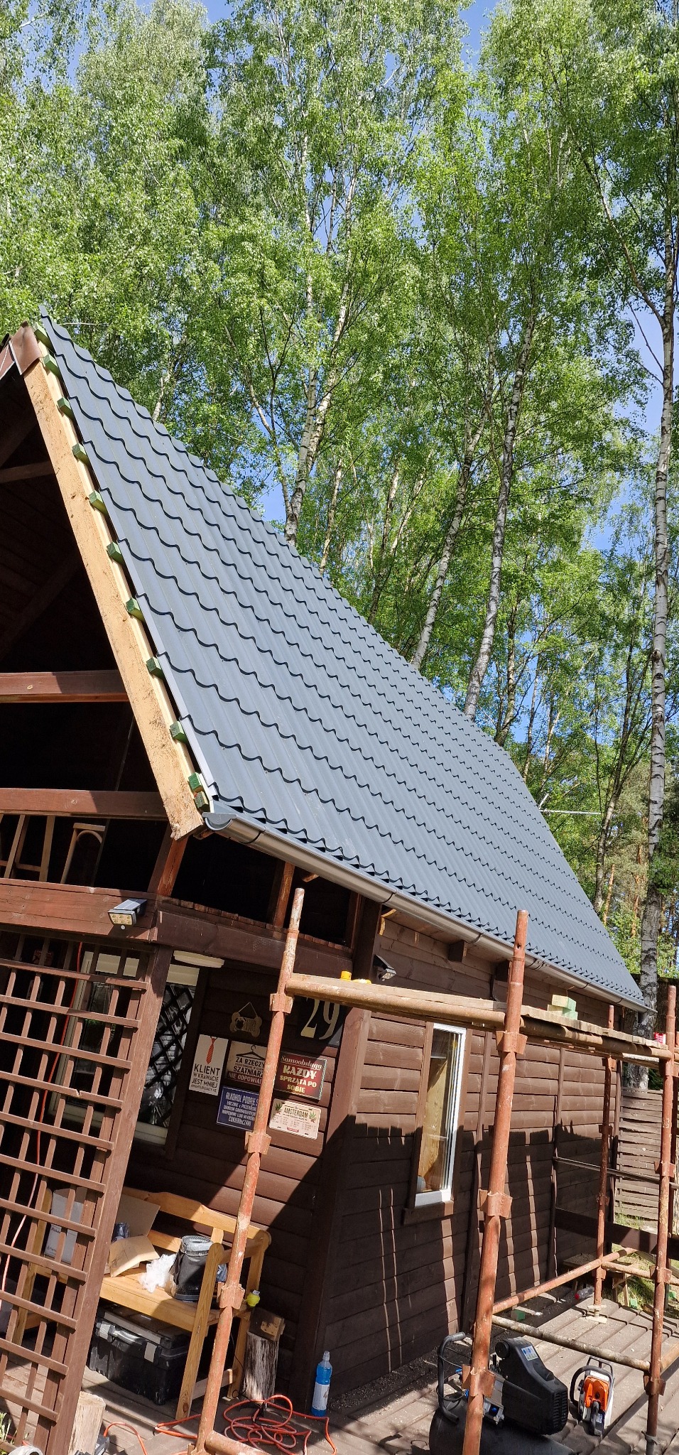 Drewniany domek z nowym, szarym dachem z blachodachówki, widoczne rusztowania i narzędzia, otoczenie leśne.