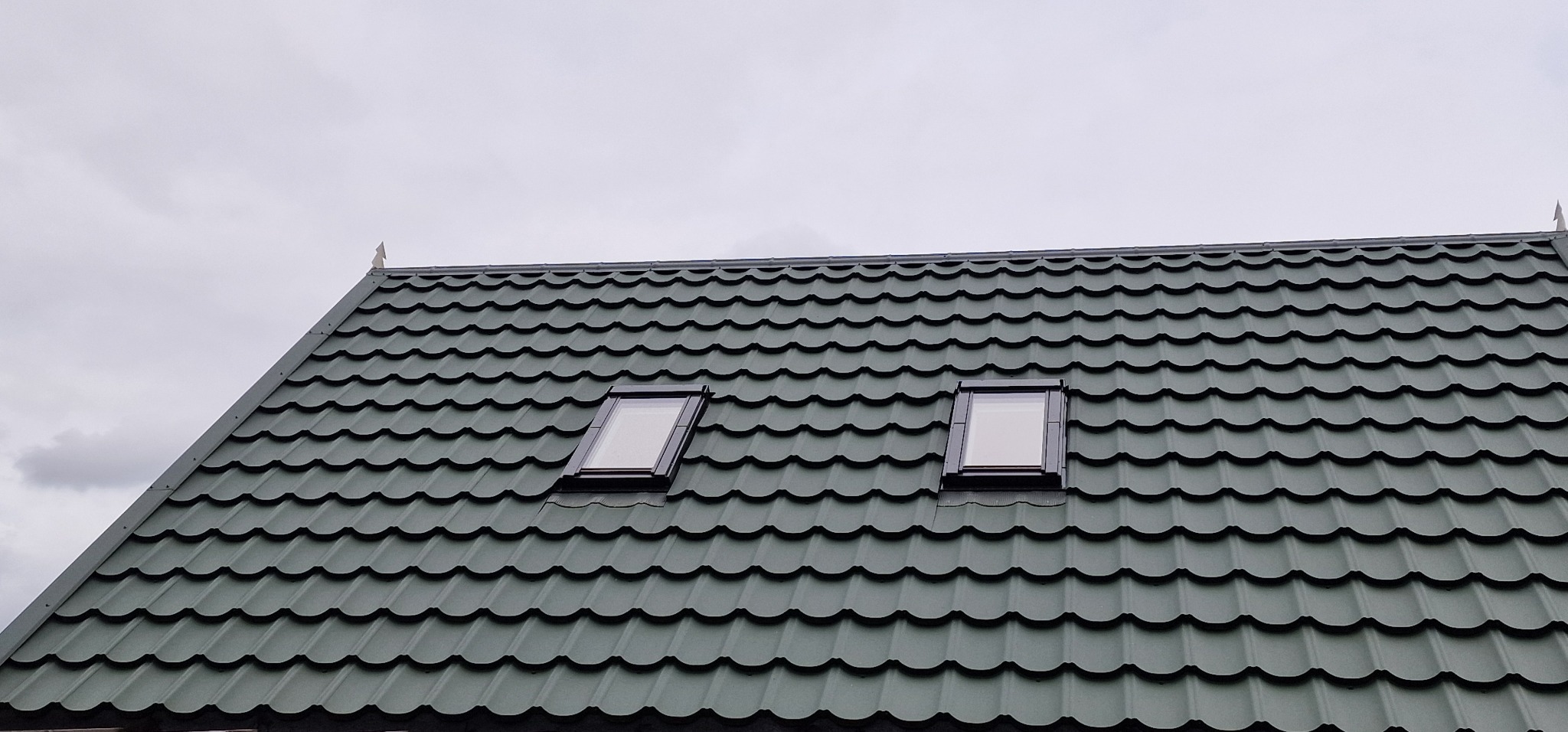 Zielony dach z blachodachówki z dwoma oknami dachowymi, widok z dołu na tle pochmurnego nieba.