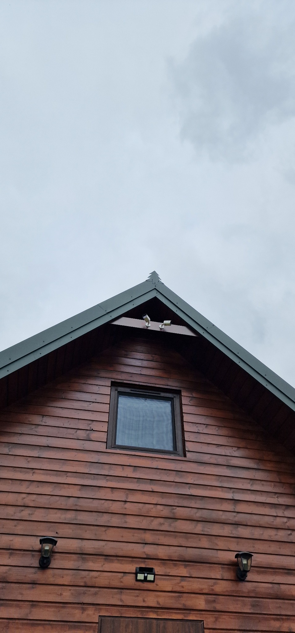 Fragment drewnianego domu z ciemnobrązową elewacją, widoczne okno z ciemną ramą, zamontowane lampy zewnętrzne i dwie kamery na szczycie dachu, pochmurne niebo w tle.