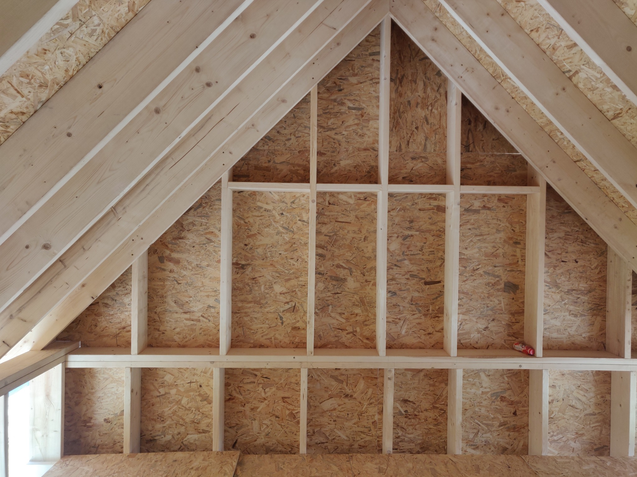 Wnętrze domu w trakcie budowy, widoczne drewniane elementy konstrukcyjne ścian i dachu, płyty OSB, częściowo widoczne okno, paczka materiałów budowlanych na półce.