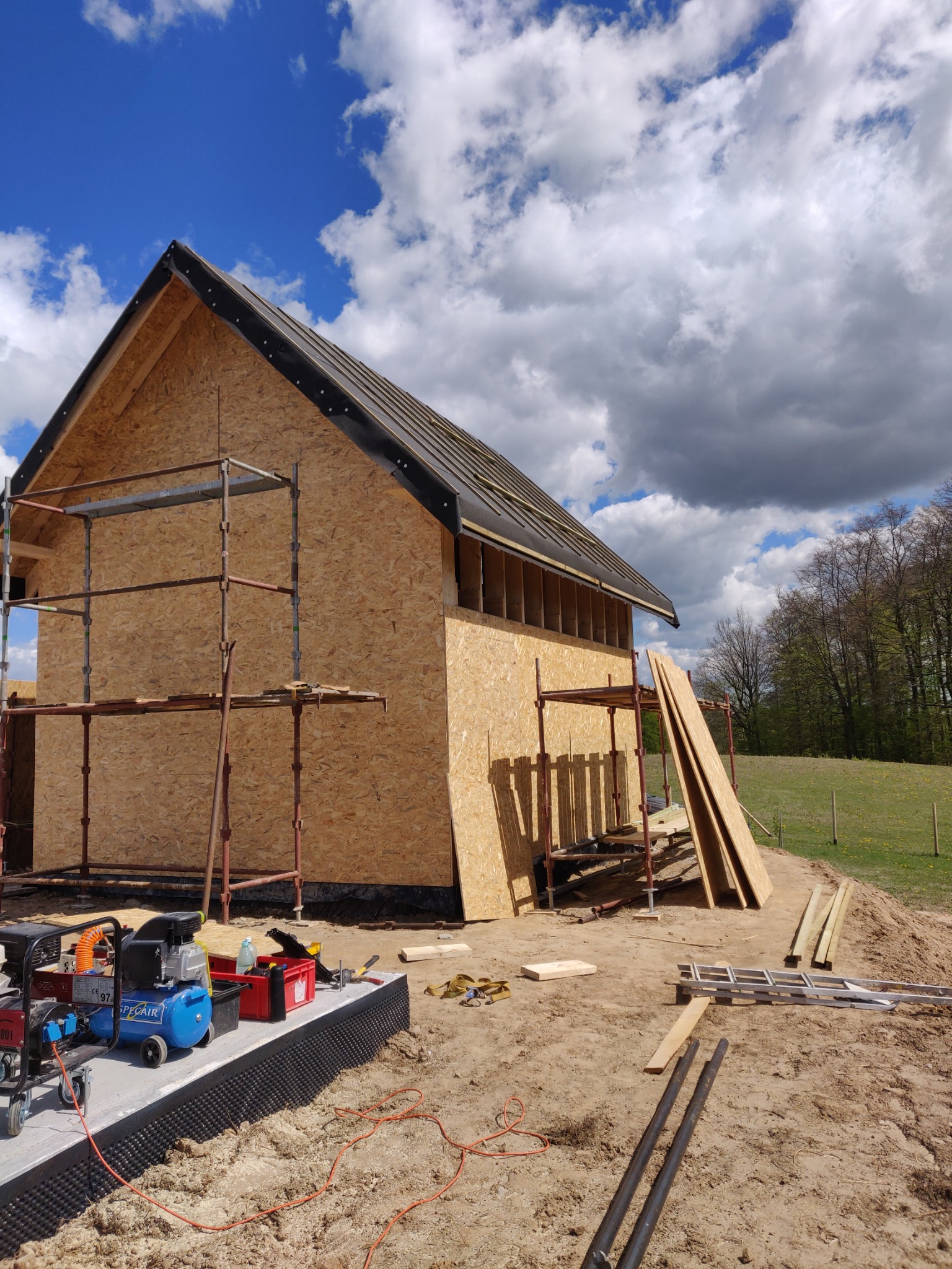 Budowa domu szkieletowego w trakcie: widoczne ściany z płyt OSB, rusztowania, częściowo wykończony dach z membraną, narzędzia i materiały budowlane na placu budowy, w tle zielona łąka i drzewa...
