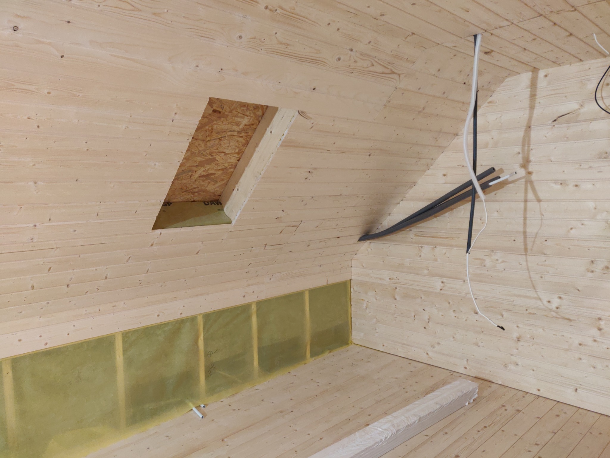 Wnętrze domu w konstrukcji szkieletowej z widoczną izolacją, instalacjami i otworem w ścianie, drewniane wykończenie ścian i podłogi.