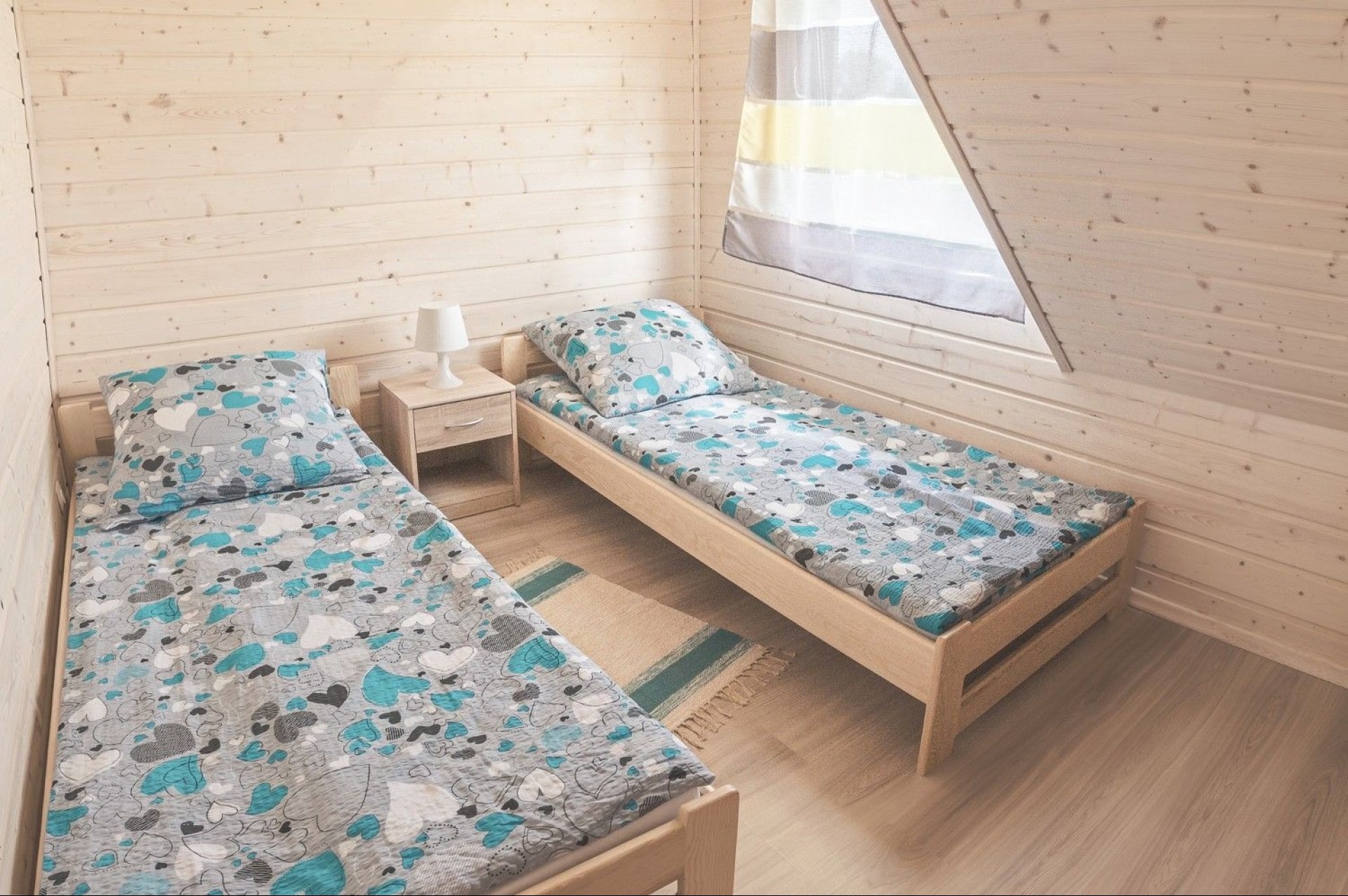 Jasne wnętrze pokoju w domu szkieletowym z dwoma pojedynczymi łóżkami, drewnianą podłogą i ścianami z desek, oświetlone naturalnym światłem z okna dachowego.