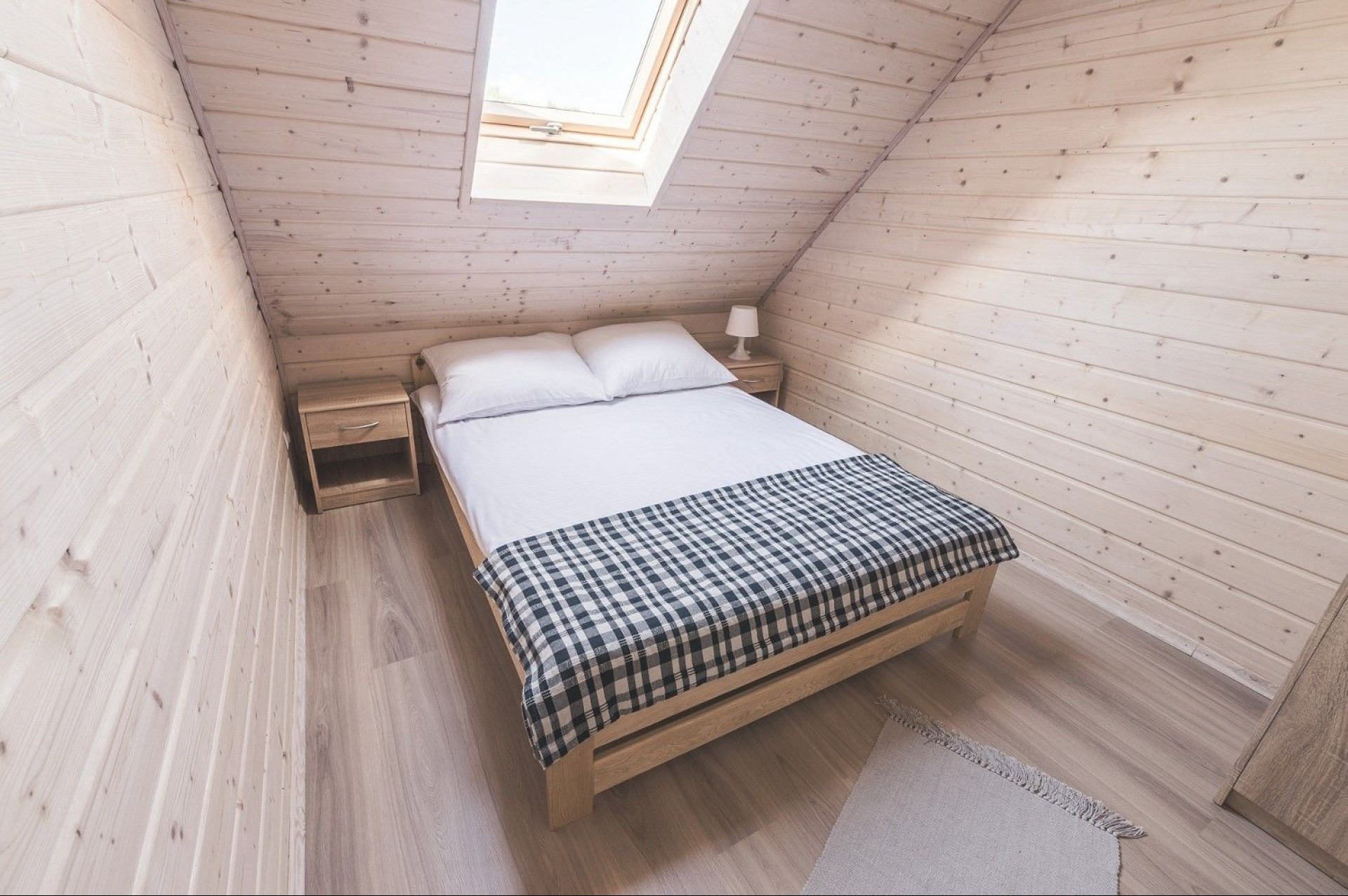Jasna sypialnia na poddaszu wykończona boazerią, z oknem dachowym, drewnianym łóżkiem przykrytym białą pościelą i pledem w kratę, oraz szafkami nocnymi po bokach.