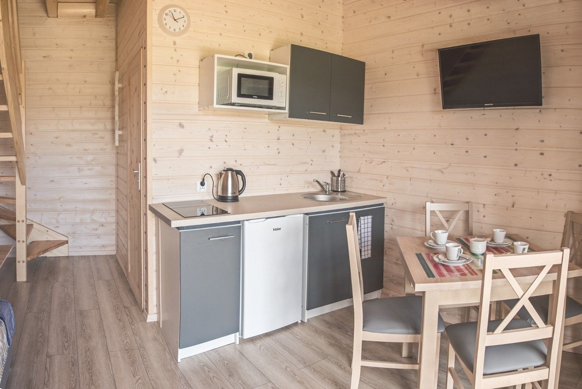 Wnętrze domku z drewnianą boazerią: aneks kuchenny z szarymi szafkami, białą lodówką, czajnikiem, mikrofalówką i stołem z zastawą dla czterech osób.