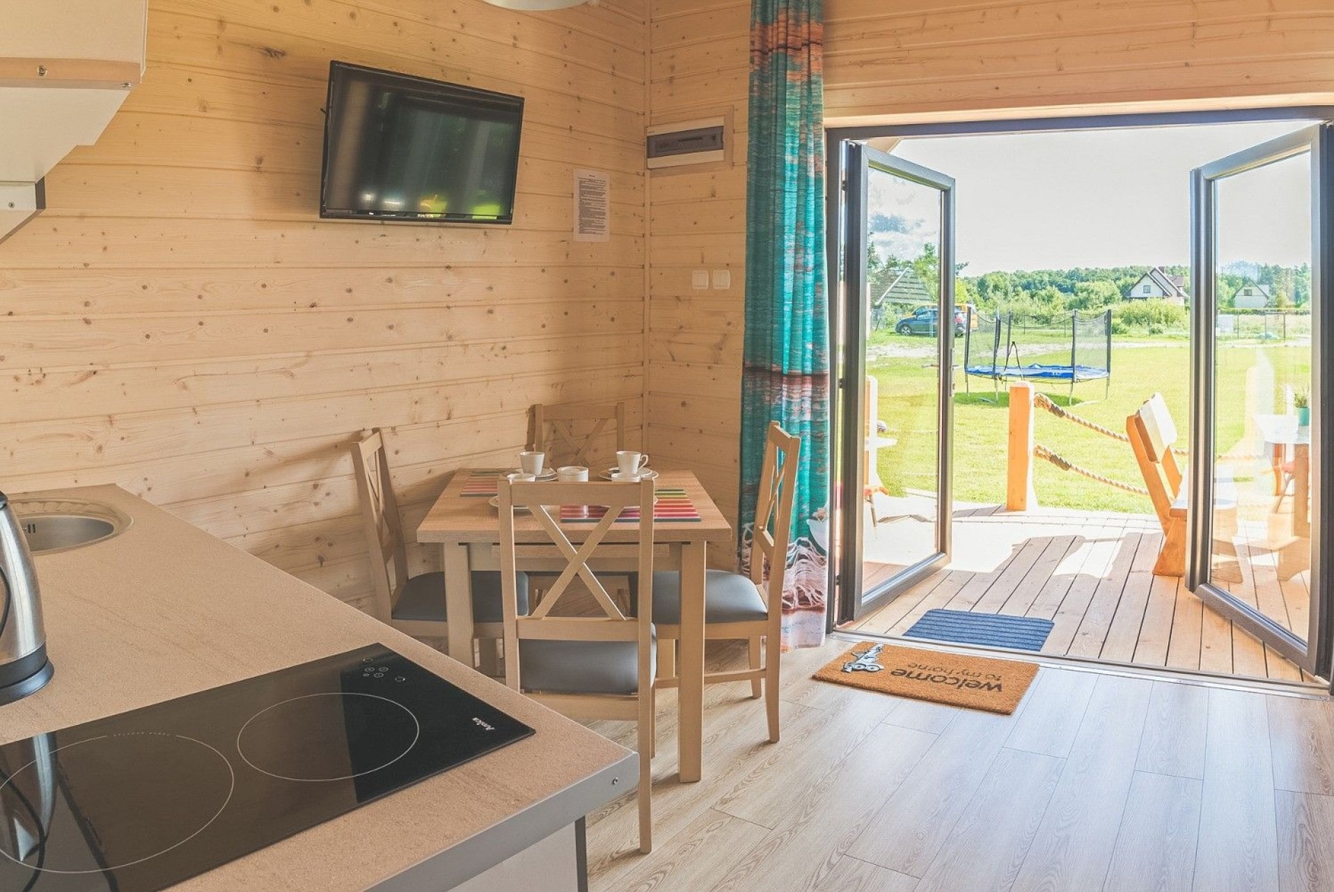 Wnętrze domku szkieletowego z drewnianymi ścianami, stołem nakrytym dla dwóch osób, aneksem kuchennym i otwartymi drzwiami na taras z widokiem na ogród z trampoliną.