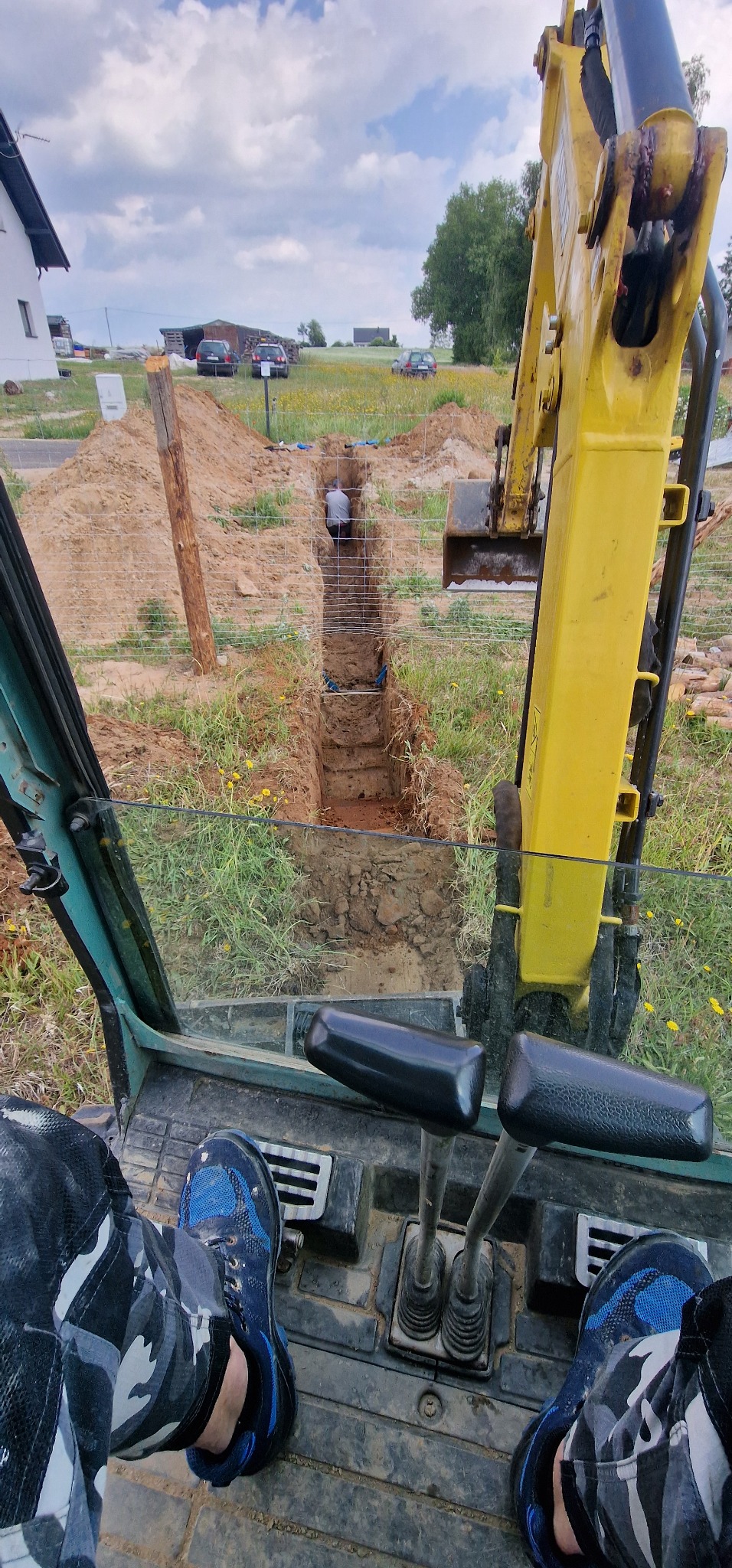 Wykop pod instalacje widziany z kabiny żółtej koparki, z widocznymi dwiema dźwigniami sterującymi i stopami operatora w niebieskich butach i spodniach moro, w tle pracownik w wykopie zabezpieczonym...