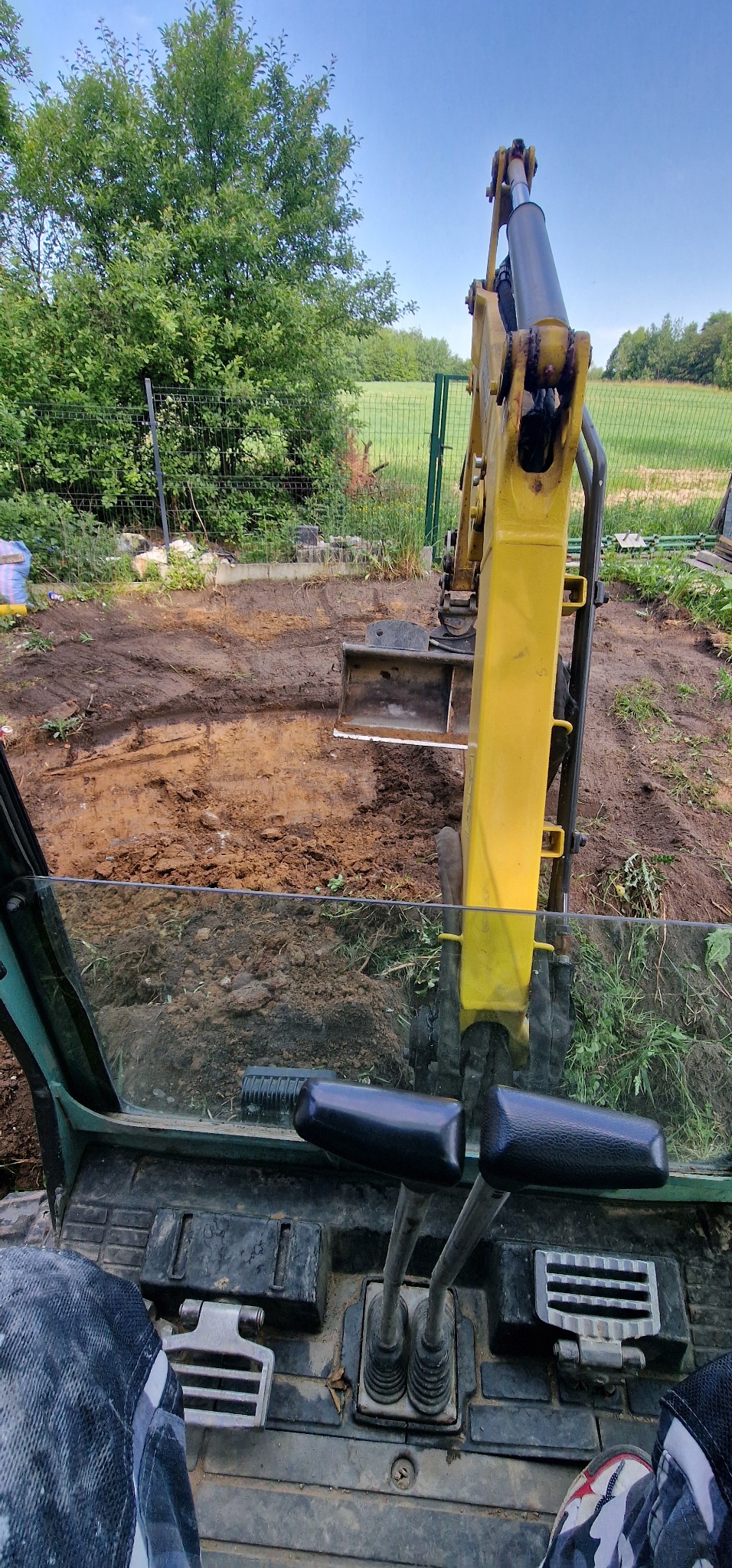 Widok z kabiny żółtej koparki na wykop w ziemi, z zielenią i ogrodzeniem w tle, pod błękitnym niebem.