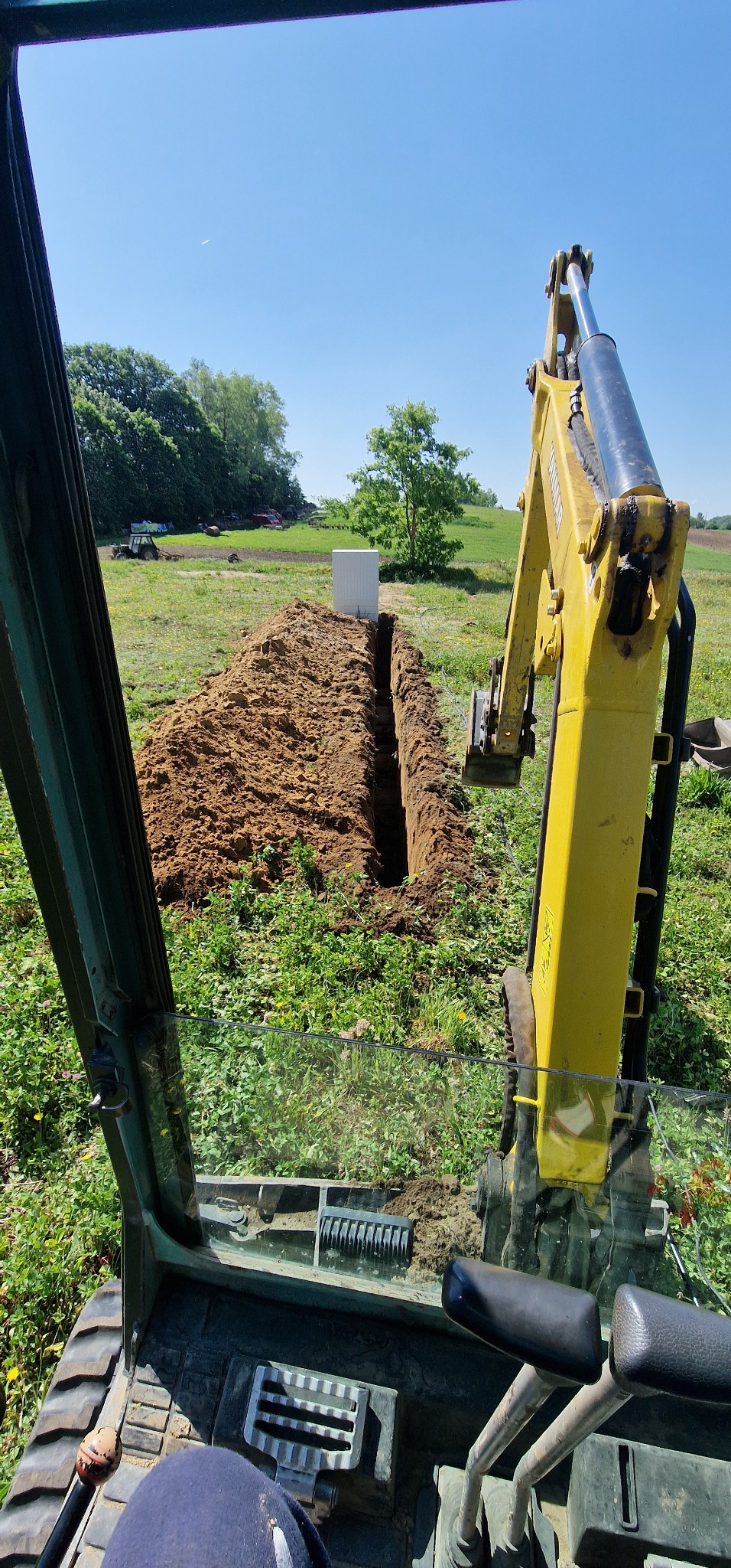 Żółta koparka widziana z kabiny podczas kopania wąskiego rowu w ziemi na zielonym polu, w tle drzewa i biały prostopadłościan