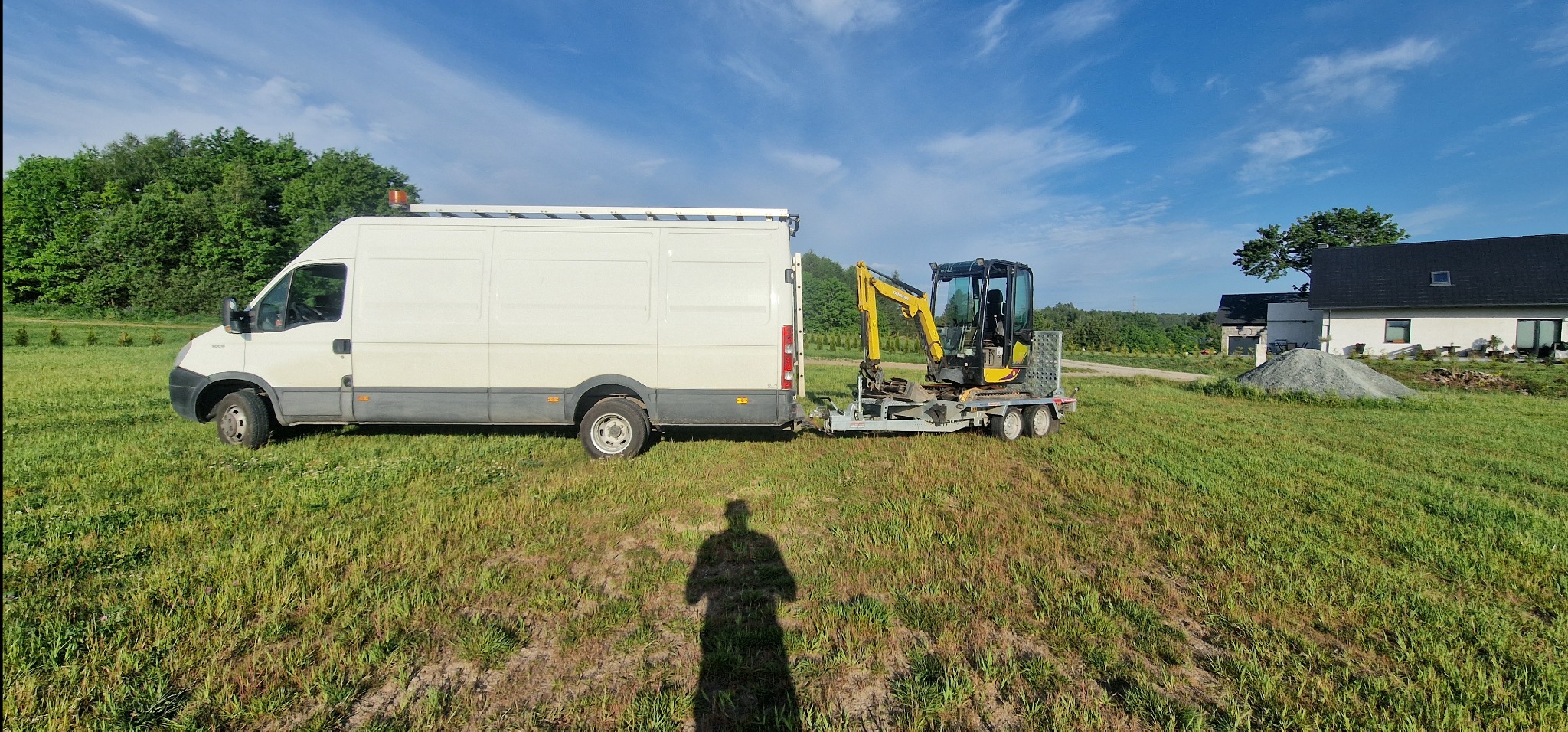 Biały bus z przyczepą i mini koparką Yanmar na łące w Wejherowie, cień fotografa na pierwszym planie, w tle domy.