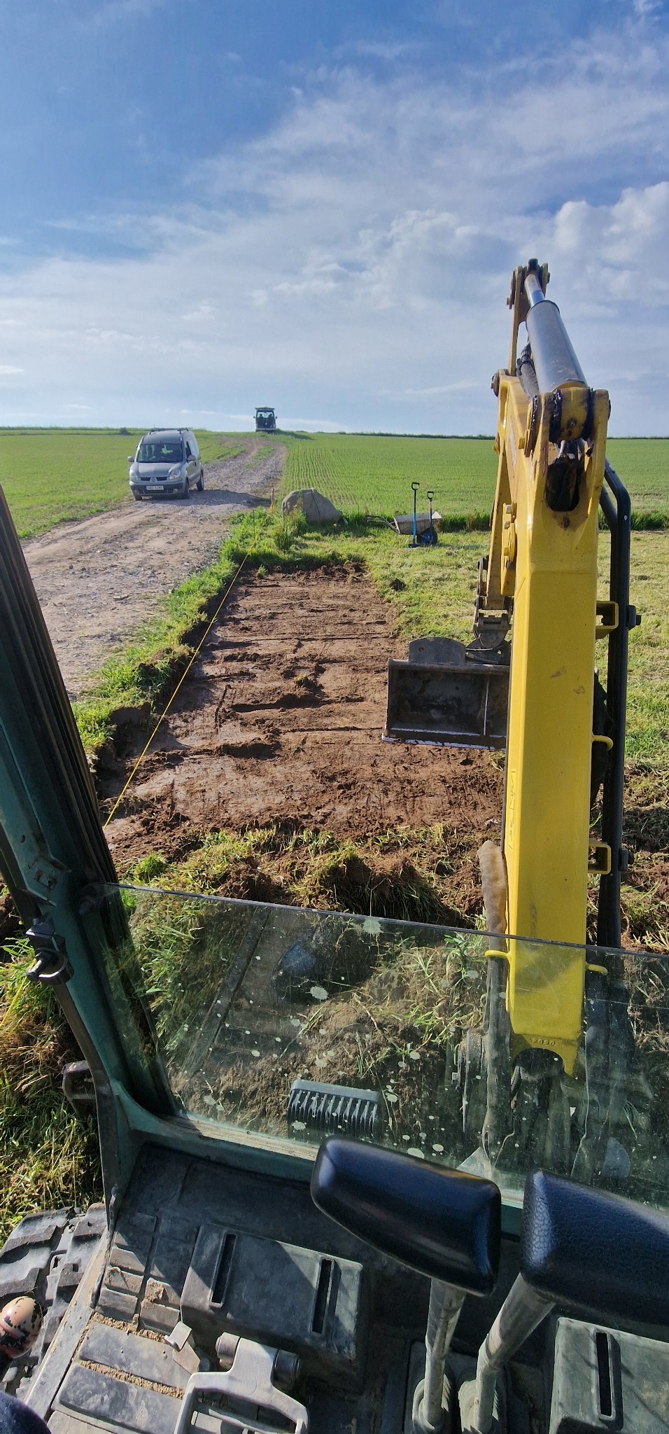 Widok z kabiny koparki na wykopany rów w ziemi, w tle pole, droga polna i dwa samochody