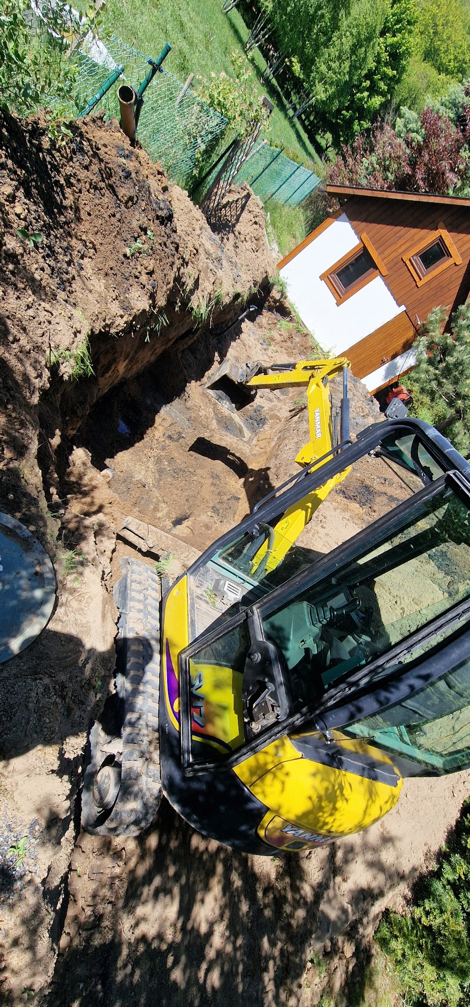 Żółta mini koparka Yanmar przy wykopie w ziemi, widok z góry na teren budowy z domkiem narzędziowym w tle, ziemia z wykopu ułożona obok, zielona trawa i drzewa w tle.