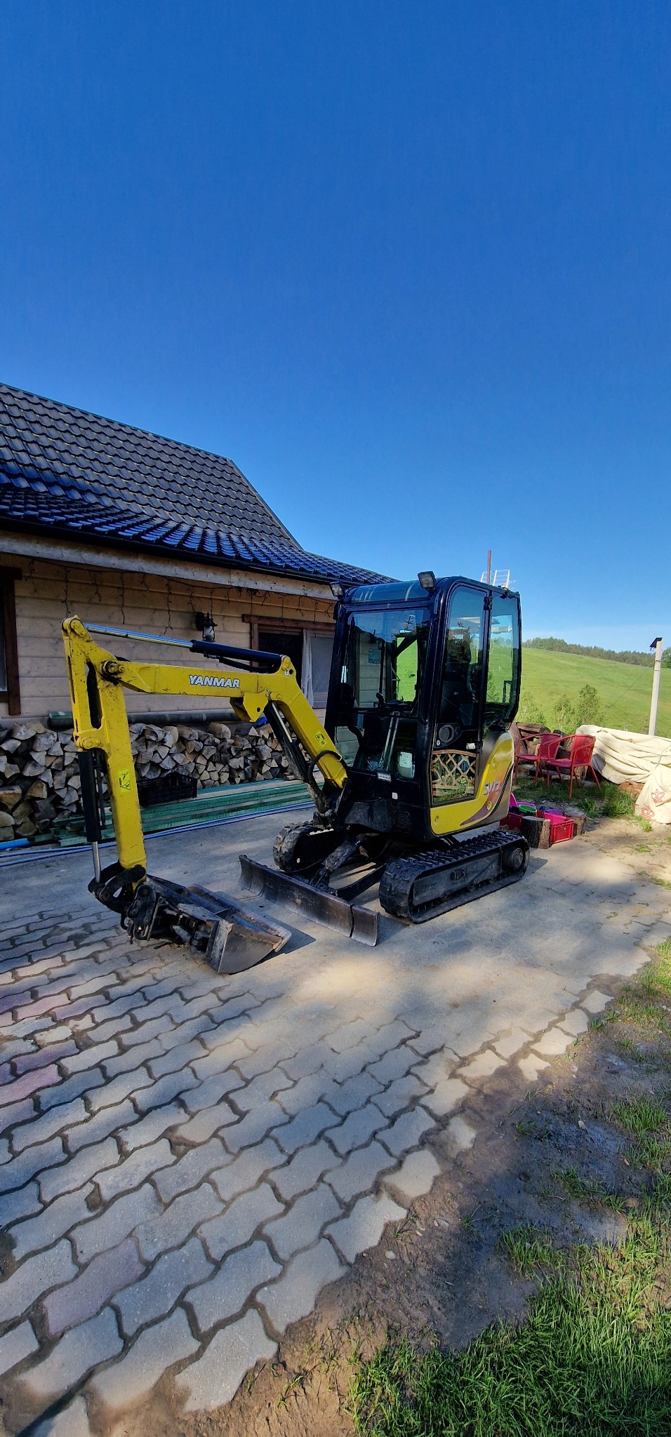 Żółta mini koparka Yanmar na kostce brukowej, z domem z ciemnym dachem i stosem drewna w tle, błękitne niebo.