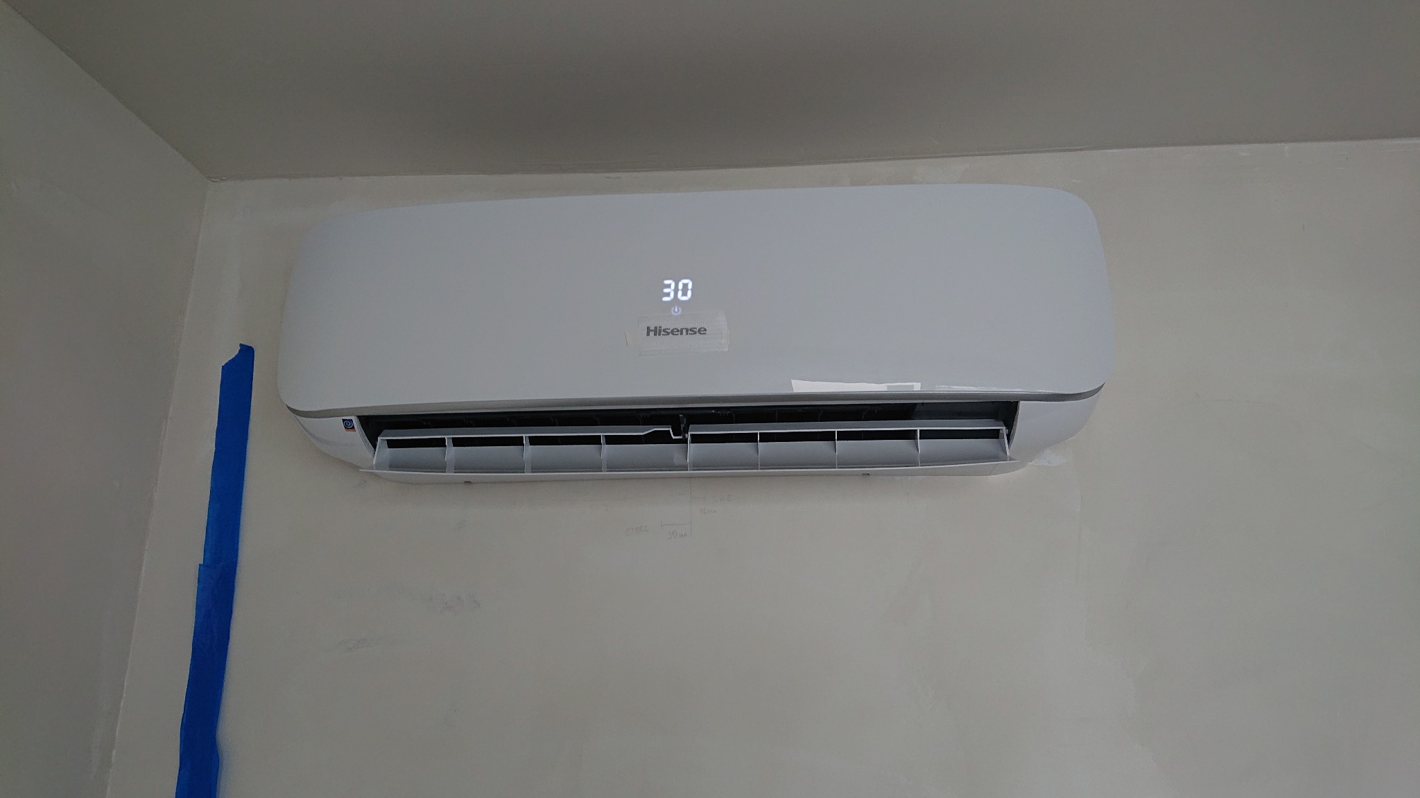 Biały klimatyzator Hisense zamontowany na ścianie z widocznymi śladami po malowaniu i niebieską taśmą malarską przy krawędzi ściany.