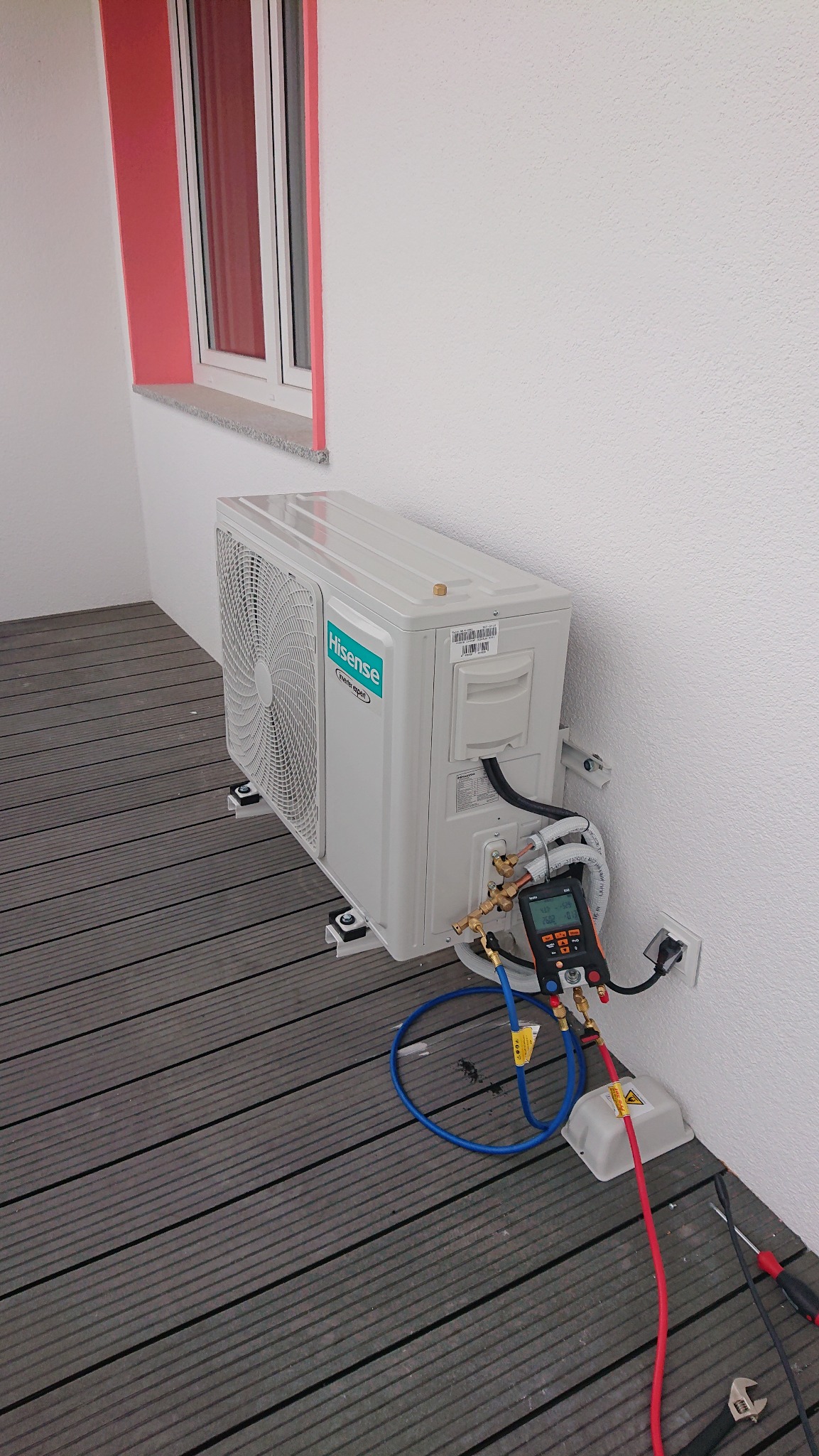 Biała jednostka zewnętrzna klimatyzacji Hisense zamontowana na wspornikach na tarasie z desek kompozytowych, podłączona do miernika ciśnienia z przewodami, obok narzędzia.
