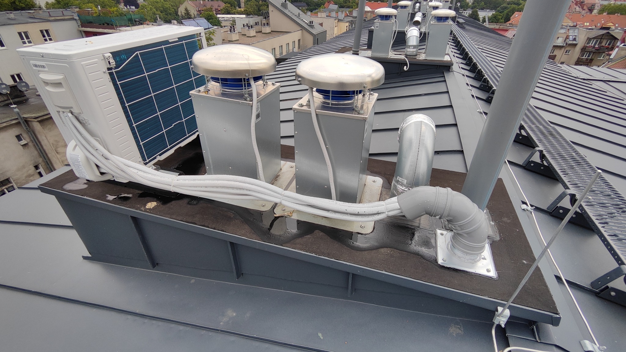 Zewnętrzna jednostka klimatyzacji i elementy wentylacji na dachu budynku, widoczne przewody i metalowe kominy wentylacyjne z okrągłymi daszkami, w tle zabudowa miejska.