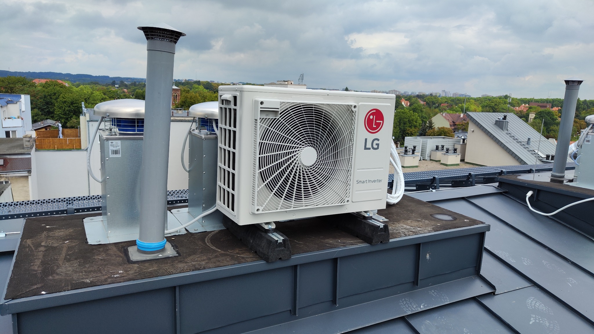 Biała jednostka zewnętrzna klimatyzacji LG Smart Inverter umieszczona na dachu budynku, widoczne przewody i elementy montażowe, w tle zabudowa miejska z zielenią.