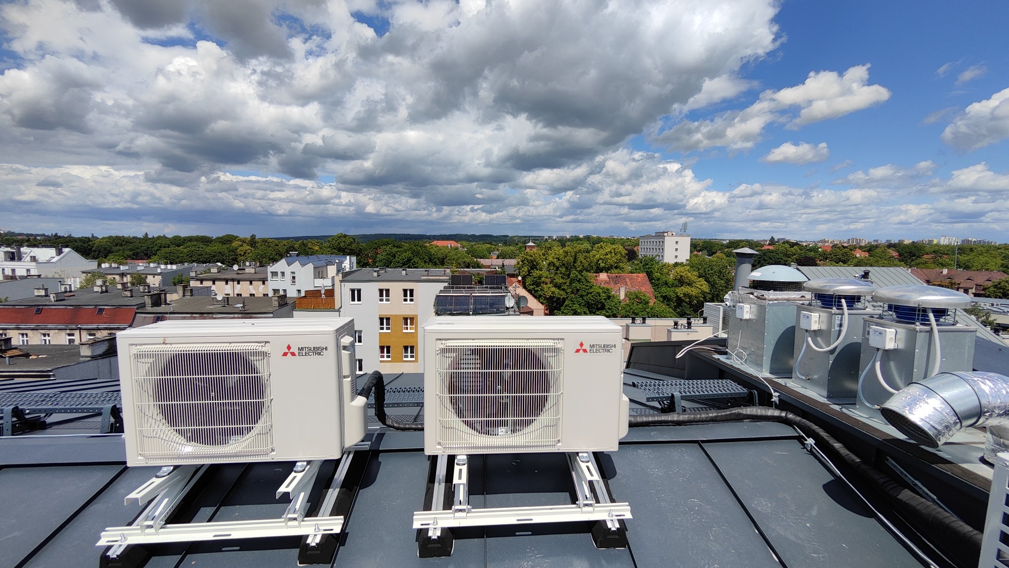 Dwa zewnętrzne moduły klimatyzacji Mitsubishi Electric zamontowane na dachu budynku w Szczecinie, widok na panoramę miasta i pochmurne niebo.