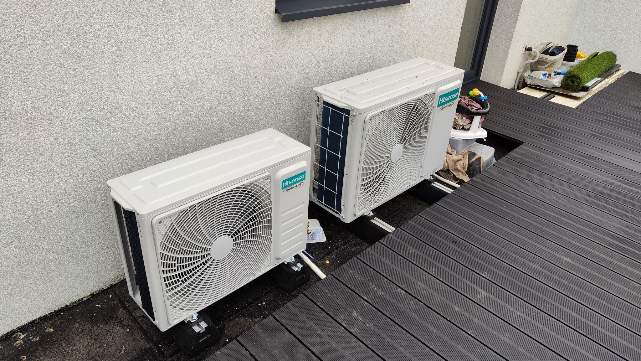 Dwa białe klimatyzatory Hisense Inverter Expert zamontowane na czarnym tarasie z desek kompozytowych, z widocznymi elementami instalacji i akcesoriami dziecięcymi w tle.