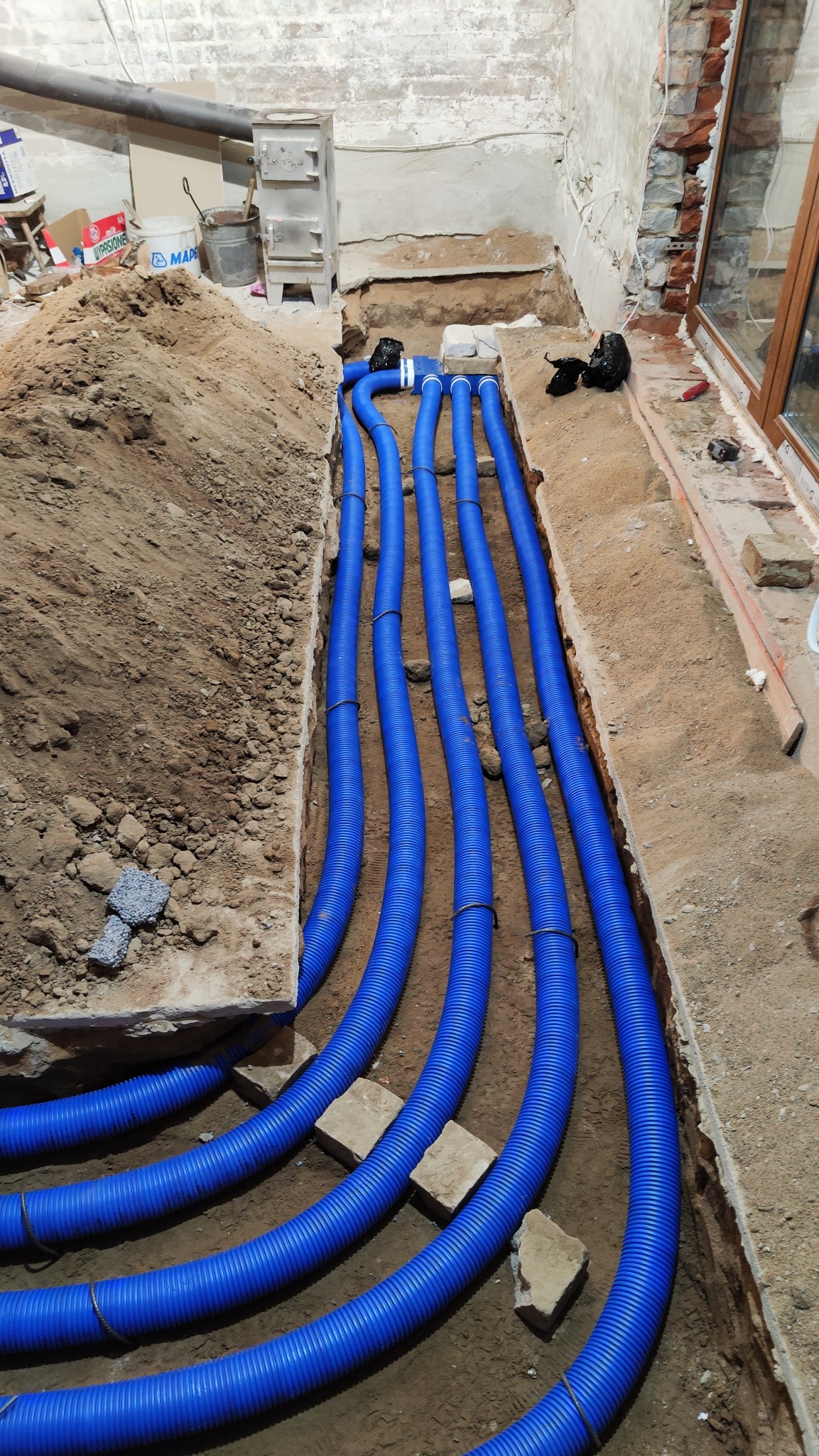 Instalacja podłogowa z niebieskich rur karbowanych w wykopie, widoczne fragmenty cegieł i ziemi, etap prac w surowym wnętrzu.