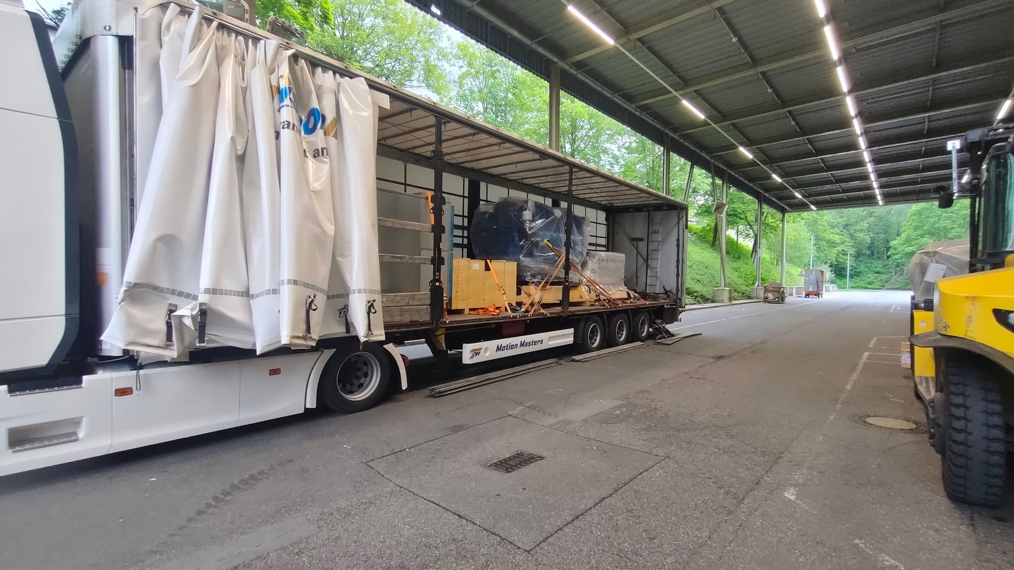 Ciężarówka z naczepą, częściowo zasłonięta plandeką, załadowana maszynami i skrzyniami, zabezpieczonymi pasami transportowymi, czeka na rozładunek. Widoczny wózek widłowy.