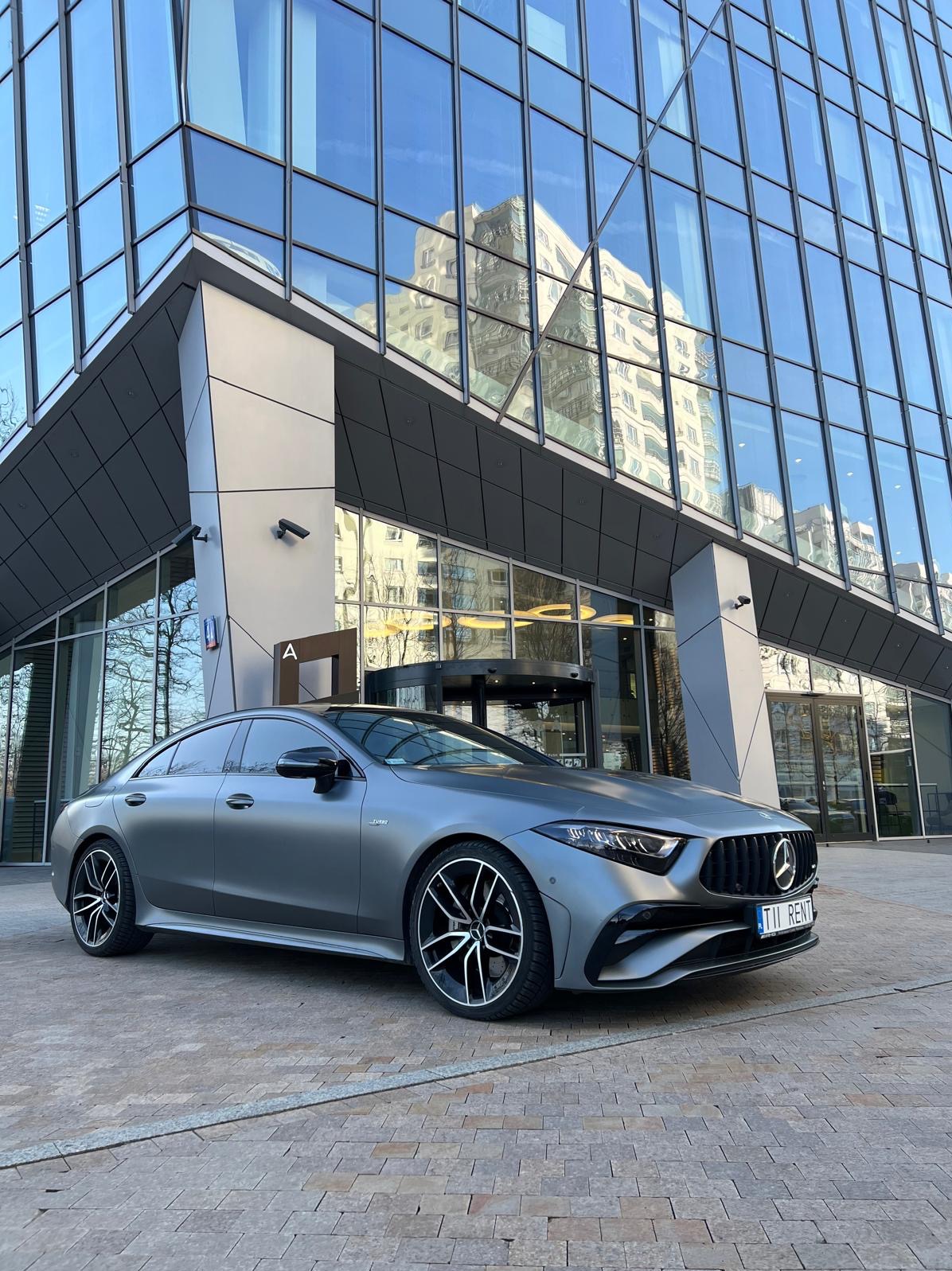 Matowy, szary Mercedes CLS z czarnymi felgami i tablicą 'TII RENT' na tle szklanego biurowca z obrotowymi drzwiami wejściowymi.