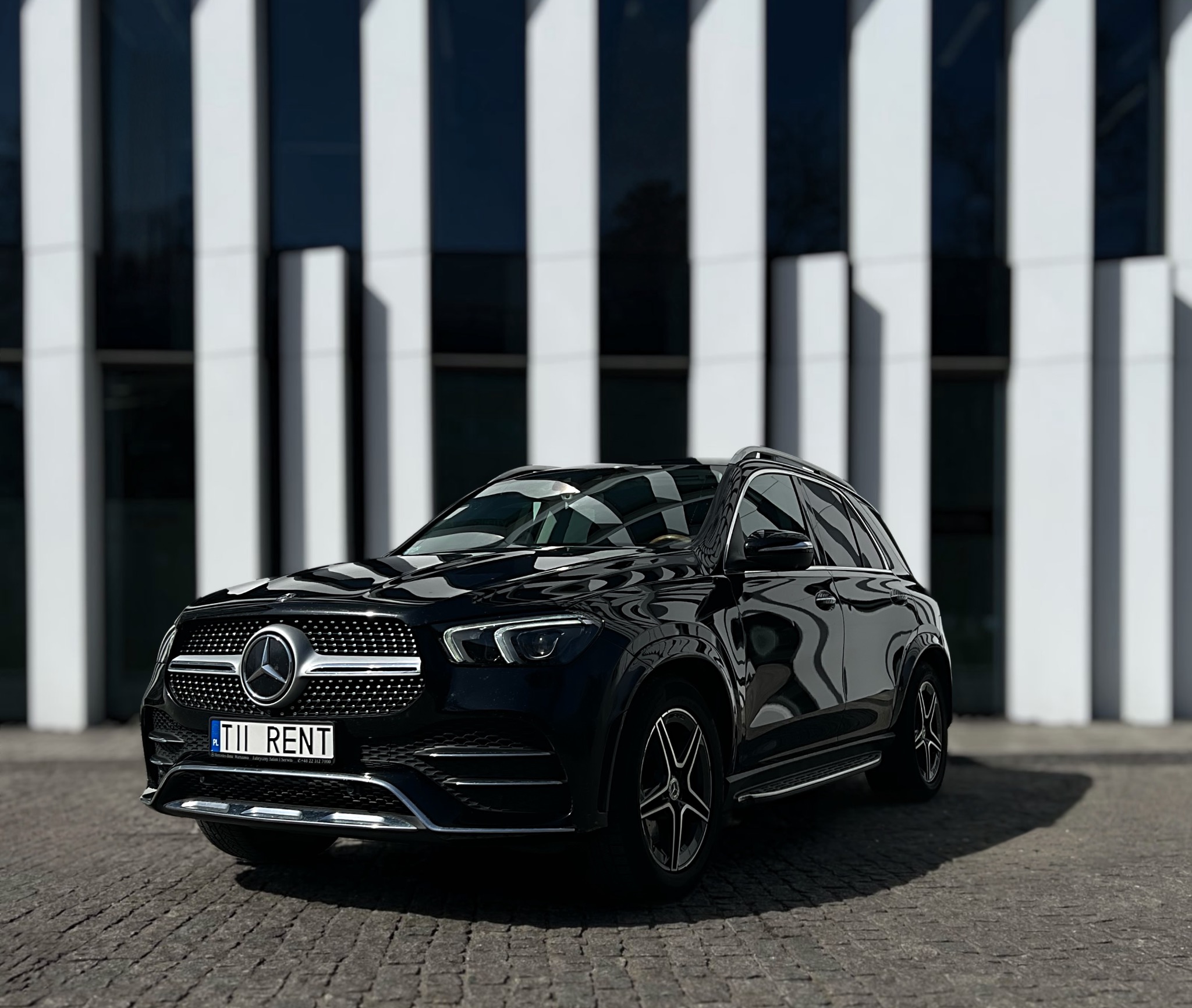 Elegancki czarny Mercedes GLE z tablicą 'TII RENT' na tle nowoczesnego budynku z białymi kolumnami. Auto na brukowanej nawierzchni, w słoneczny dzień.