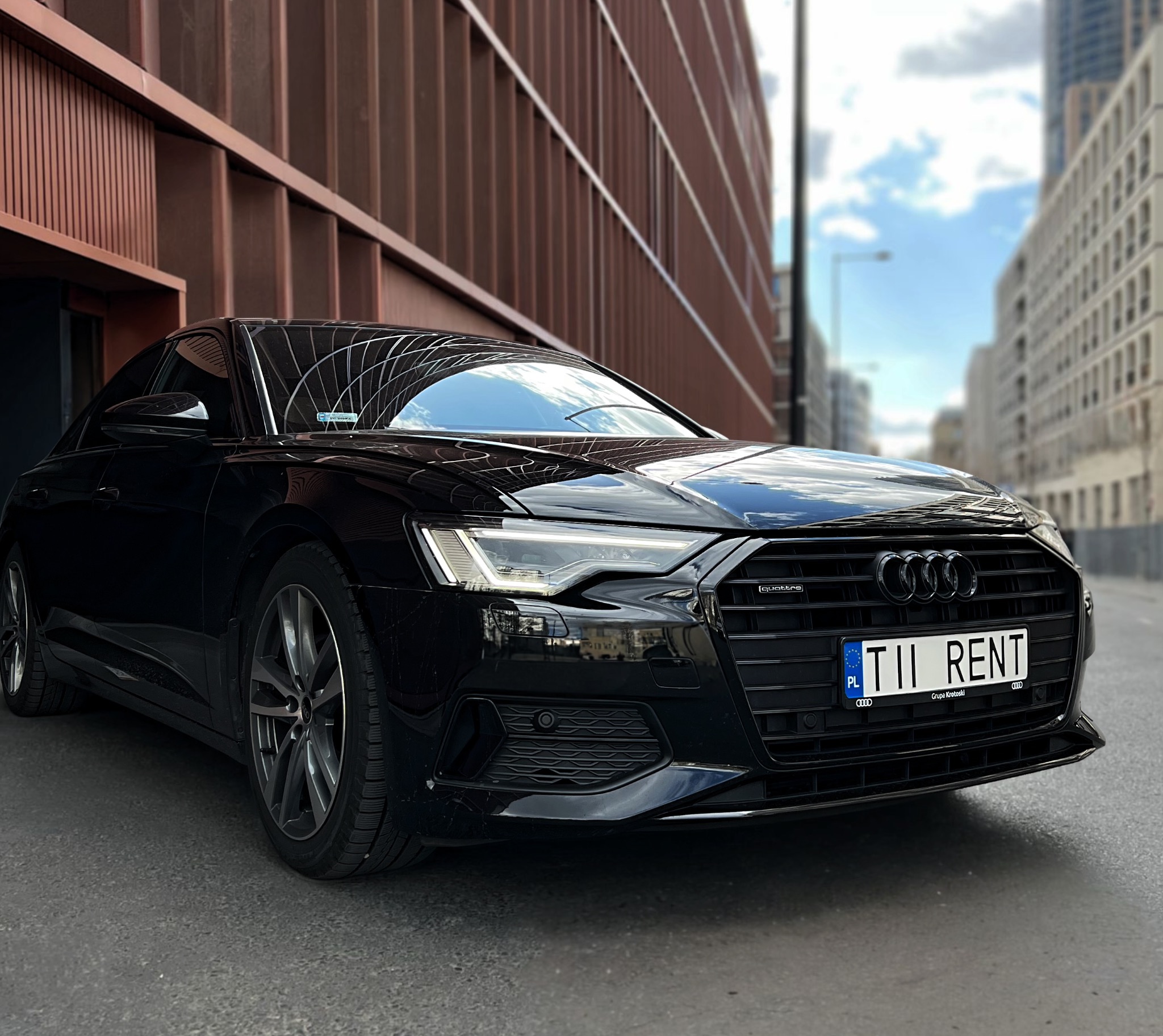 Czarny samochód Audi A6 na tle nowoczesnego budynku z brązową elewacją. Tablica rejestracyjna z napisem 'TII RENT'.