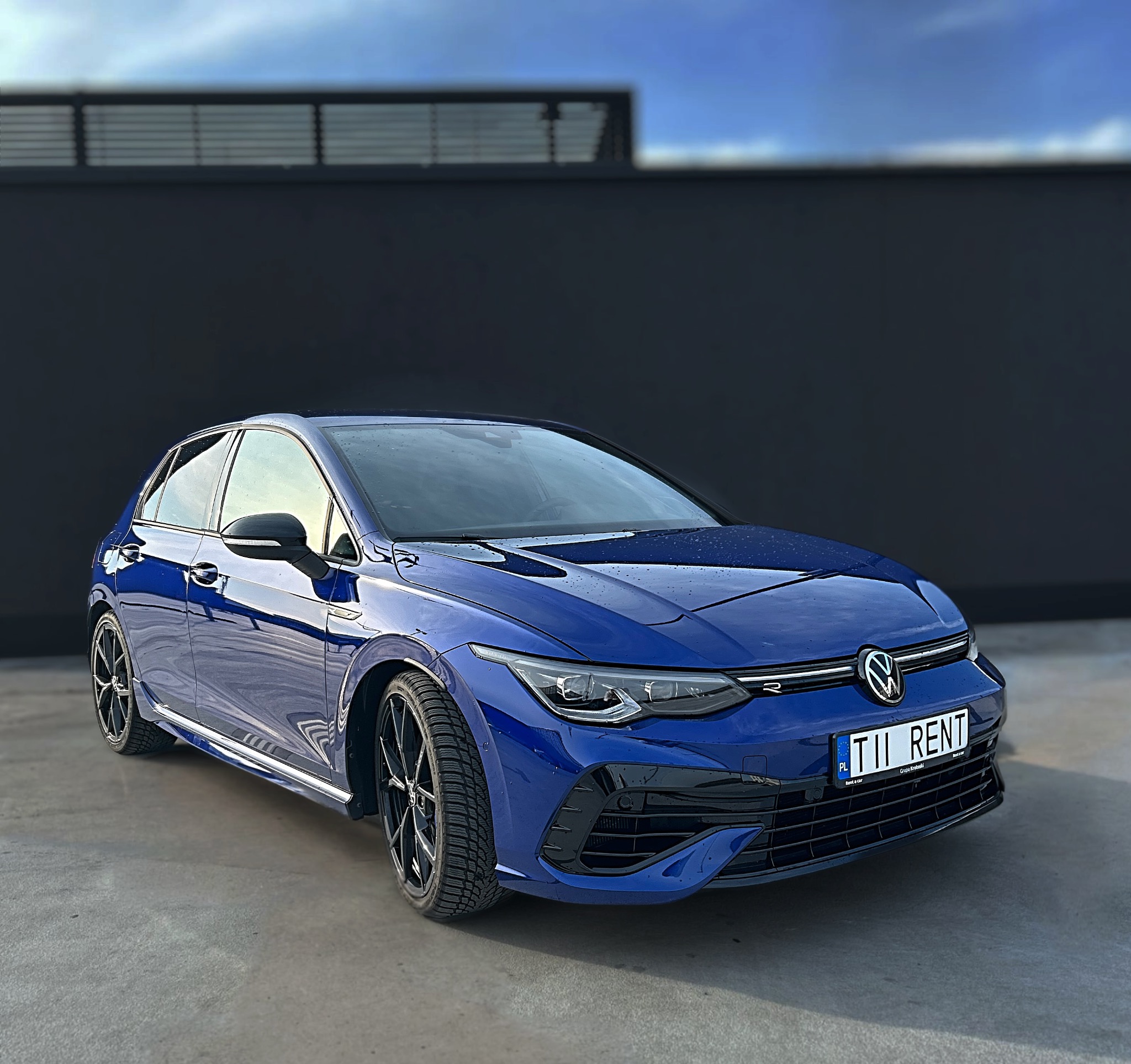 Błękitny Volkswagen Golf 8 R na tle ciemnej ściany. Widoczna tablica rejestracyjna 'TII RENT', co sugeruje wynajem auta. Nowoczesny design, sportowy wygląd.