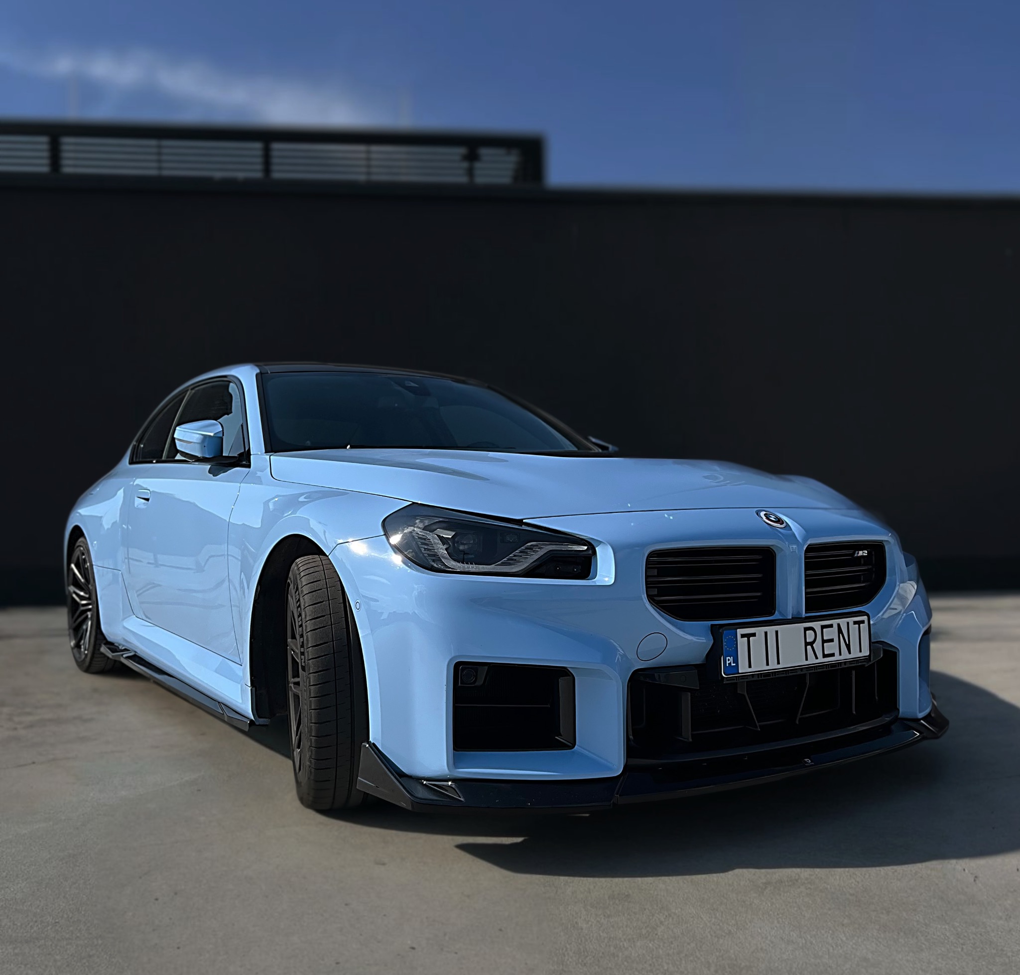Błękitny, sportowy samochód BMW M2 z czarnymi akcentami, zaparkowany na tle ciemnego muru. Tablica rejestracyjna z napisem 'RENT'.