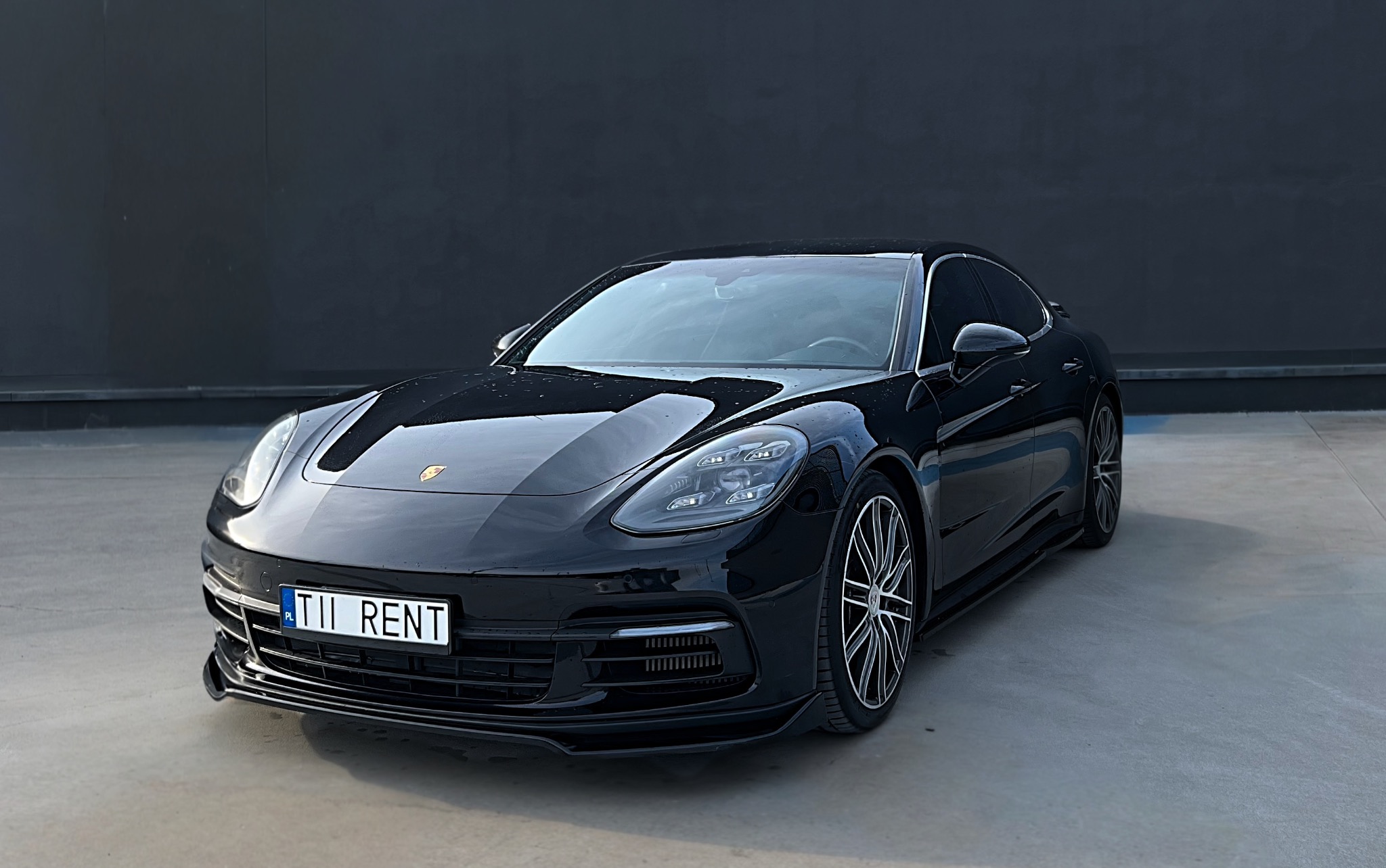 Czarna, luksusowa limuzyna Porsche Panamera, z tablicą rejestracyjną 'TII RENT', prezentowana na tle minimalistycznej, szarej ściany. Elegancki design, połysk lakieru.