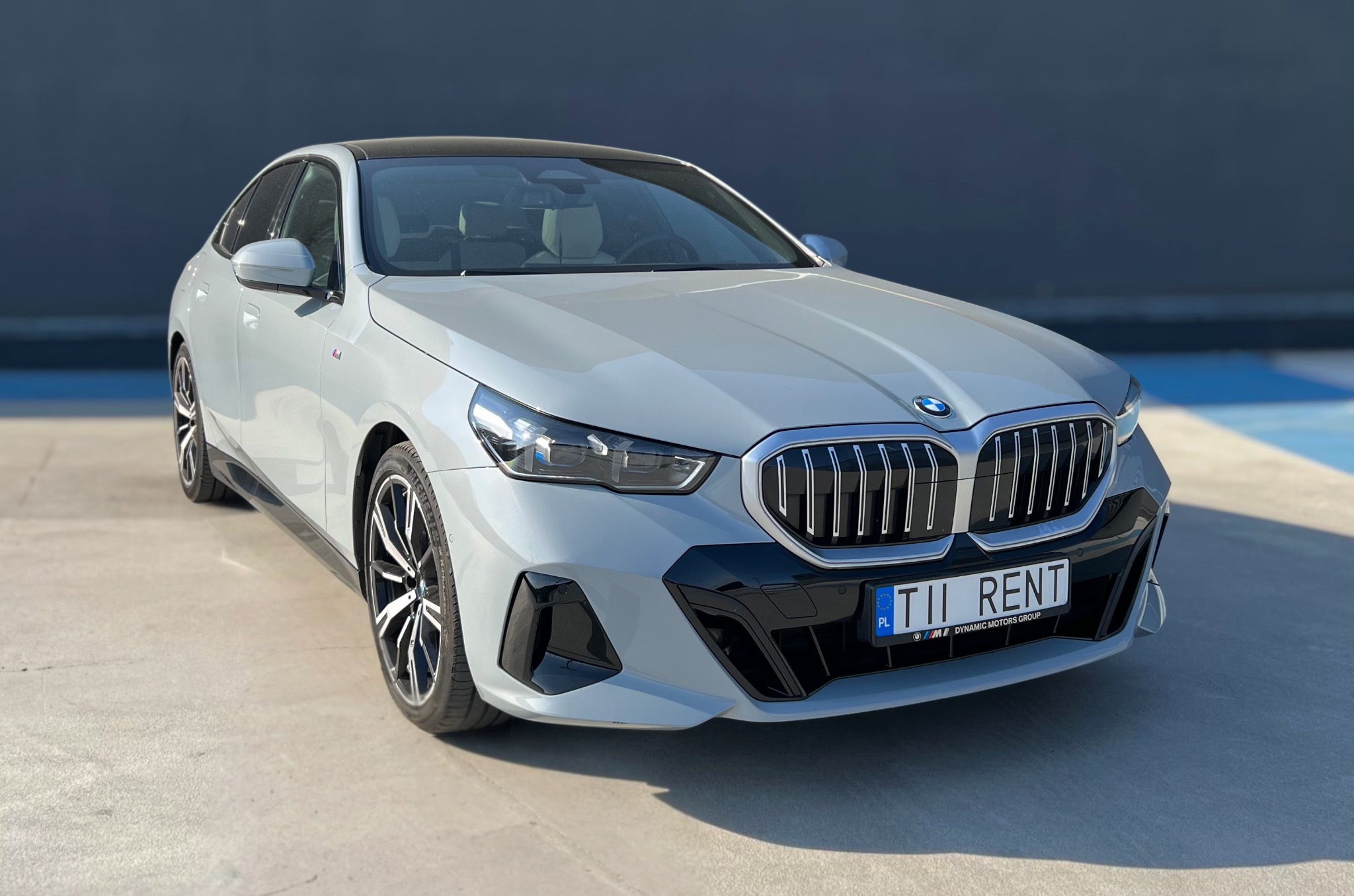 Nowoczesny, szary BMW 2 Gran Coupe z czarnym dachem, widok z przodu, tablica rejestracyjna z napisem 'RENT', na betonowym podłożu i tle ciemnej ściany.