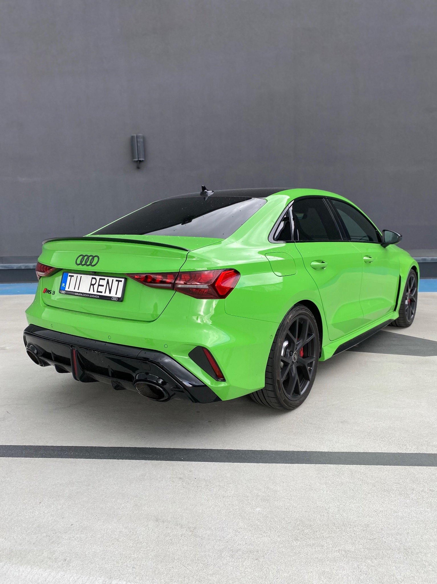 Jaskrawozielony Audi RS3 z czarnymi felgami na tle szarej ściany. Tablica rejestracyjna z napisem 'TII RENT'.