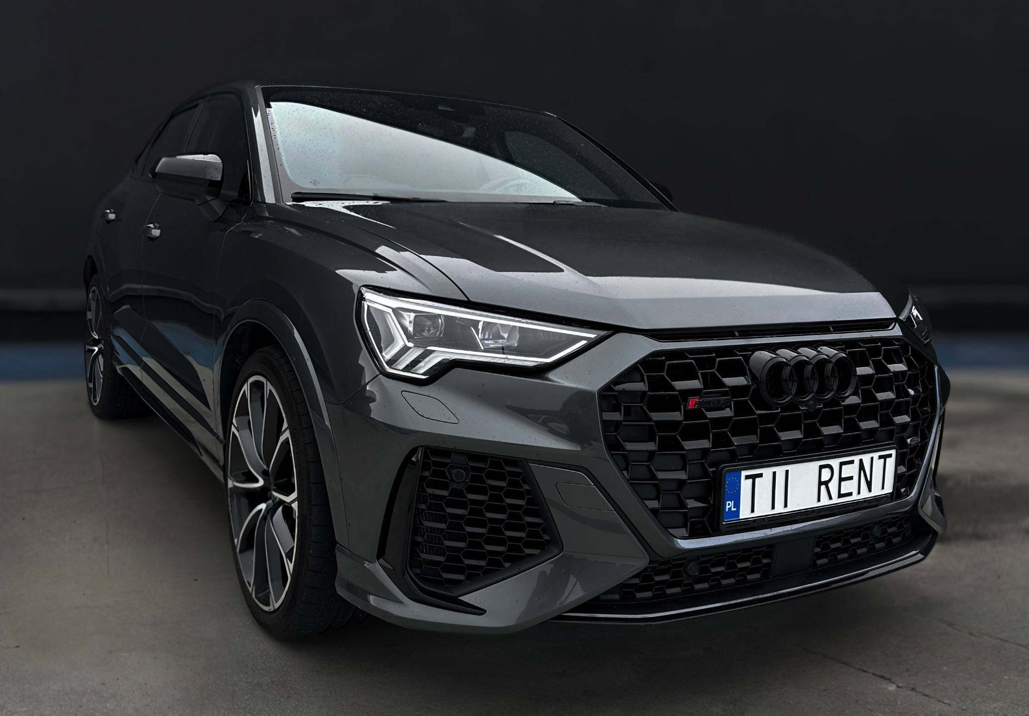 Grafitowy Audi RS Q3 z tablicą rejestracyjną 'TII RENT' na betonowym podłożu, z rozmytym, ciemnym tłem. Widok z przodu, lekko z lewej strony, z uwzględnieniem detali grilla i reflektorów.
