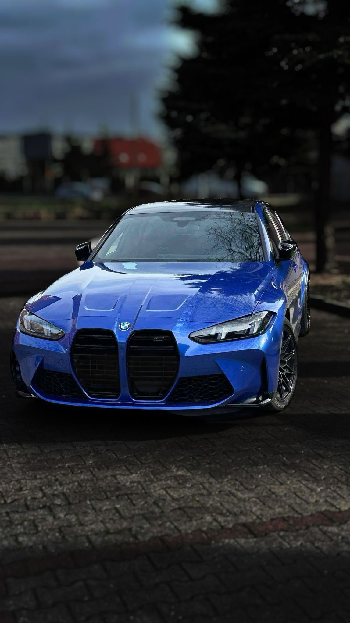 Błękitne BMW M4 z czarnym dachem, zaparkowane na brukowanej nawierzchni. Widok z przodu, z lekkim kątem. W tle drzewo i budynek. Oświetlenie naturalne, pochmurny dzień.