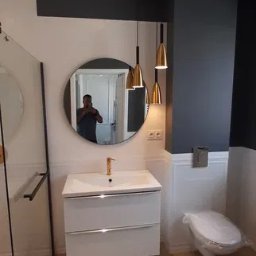 DIMBUD PATRYK JAKUBOWSKI - Wykończona łazienka z parkietem ułożonym w jodełkę, białą szafką pod umywalką z złotą baterią, okrągłym lustrem, wiszącymi złotymi lampami i toaletą owiniętą folią ochronną.