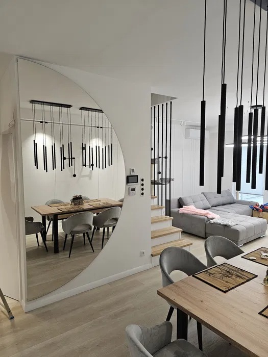 Nowoczesne wnętrze: Lustro w kształcie łuku odbija stół jadalny z szarymi krzesłami, wiszące czarne lampy, drewniane schody i szarą sofę z różowym pledem.