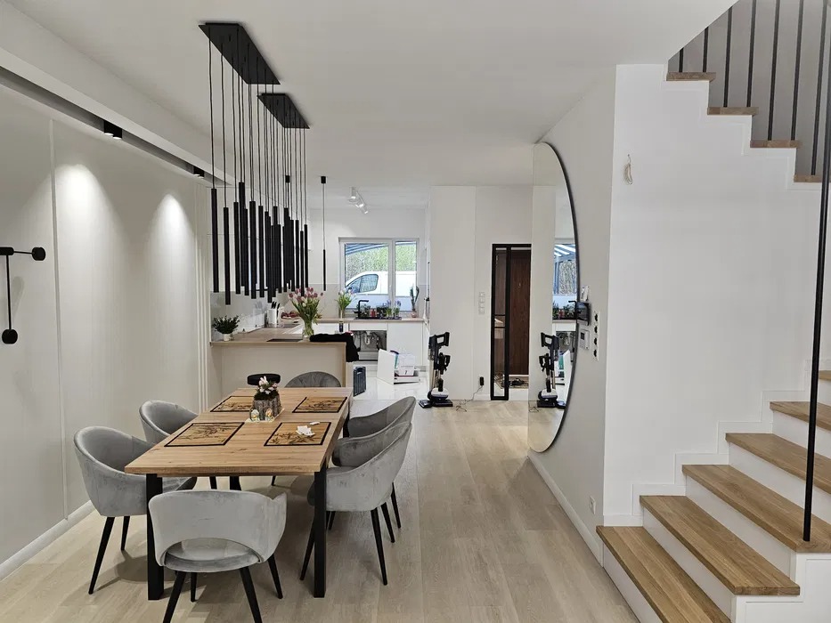 Jasne wnętrze domu z drewnianym stołem jadalnym, szarymi krzesłami, wiszącymi czarnymi lampami i schodami z drewnianymi stopniami, widok na otwartą kuchnię w tle.